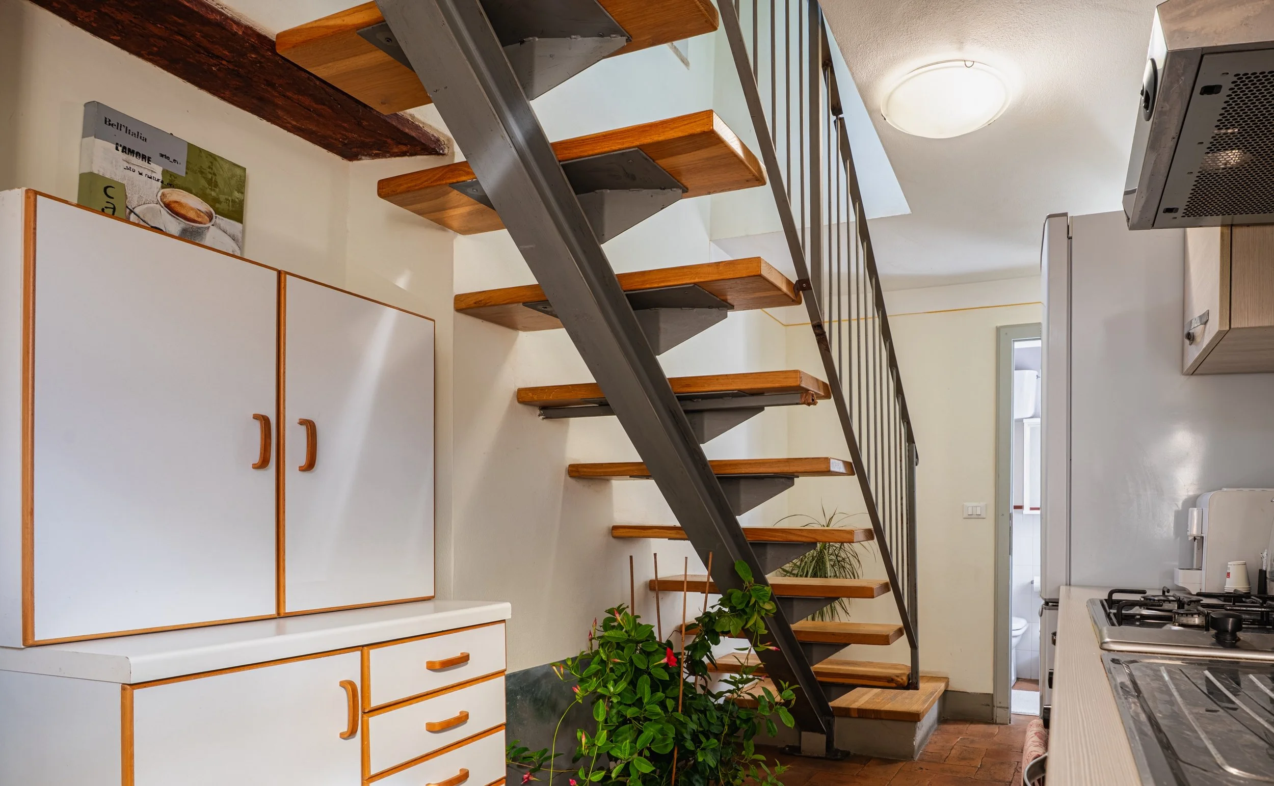 Interno di una cucina con scala di legno e metallo, mobile bianco con maniglie in legno, piante, e accessori per cucinare.