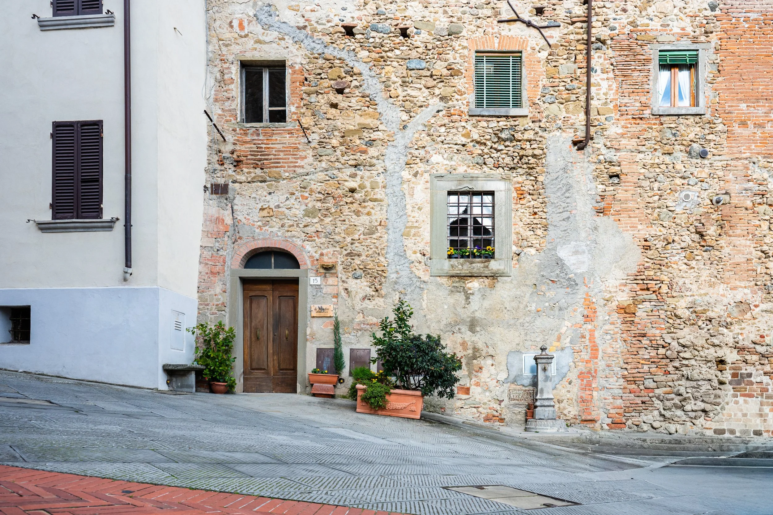 Veduta di una facciata di un edificio con mura in pietra e mattoni, finestra con inferriata e fiori, porta di legno e piante in vaso.