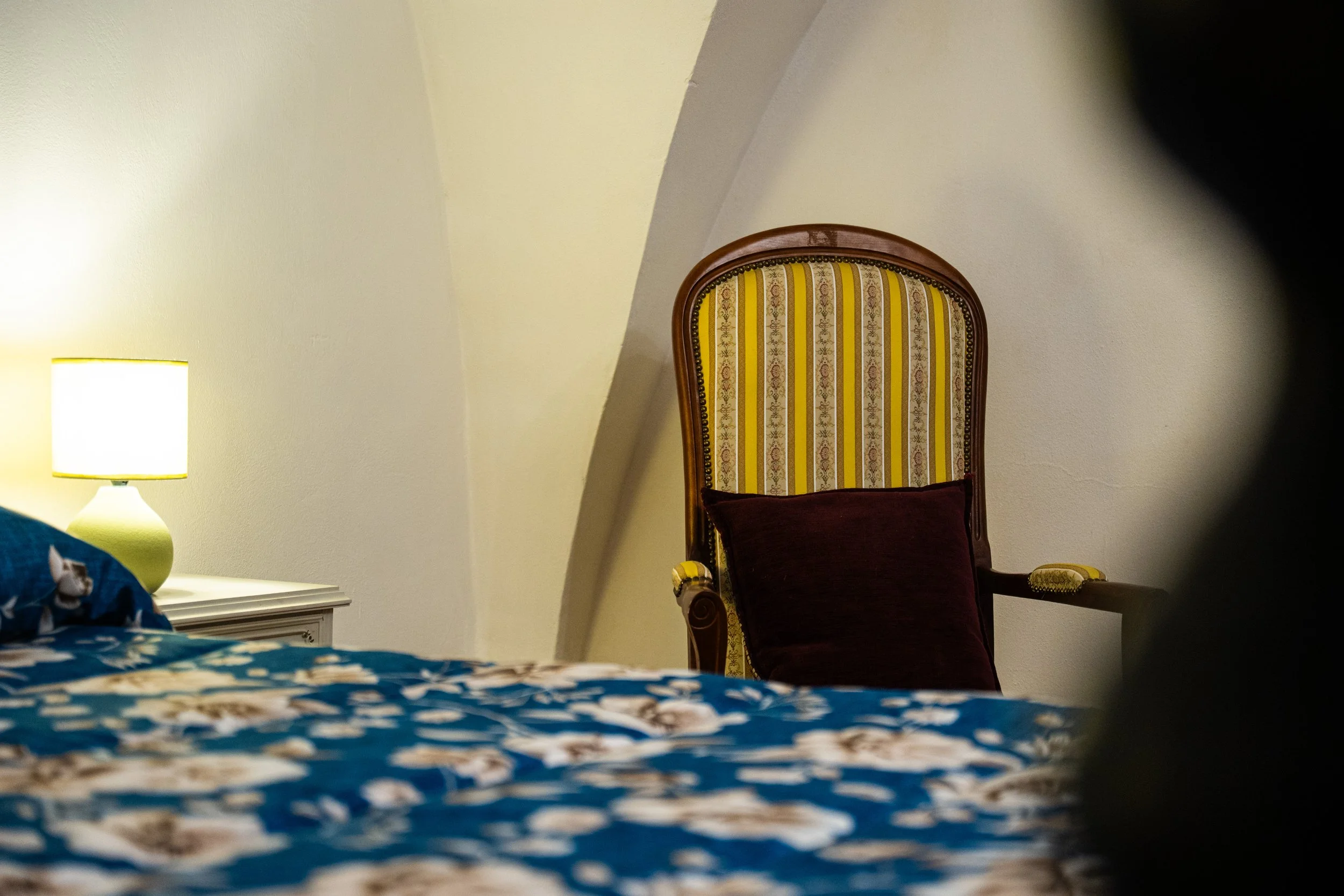 Una stanza da letto con un comodino bianco, una lampada accesa, un letto con biancheria blu con motivi floreali e un'appendiabiti in legno con uno sgabello e un cuscino viola, una sedia con schienale imbottito con tessuto a righe colorate e dettagli 