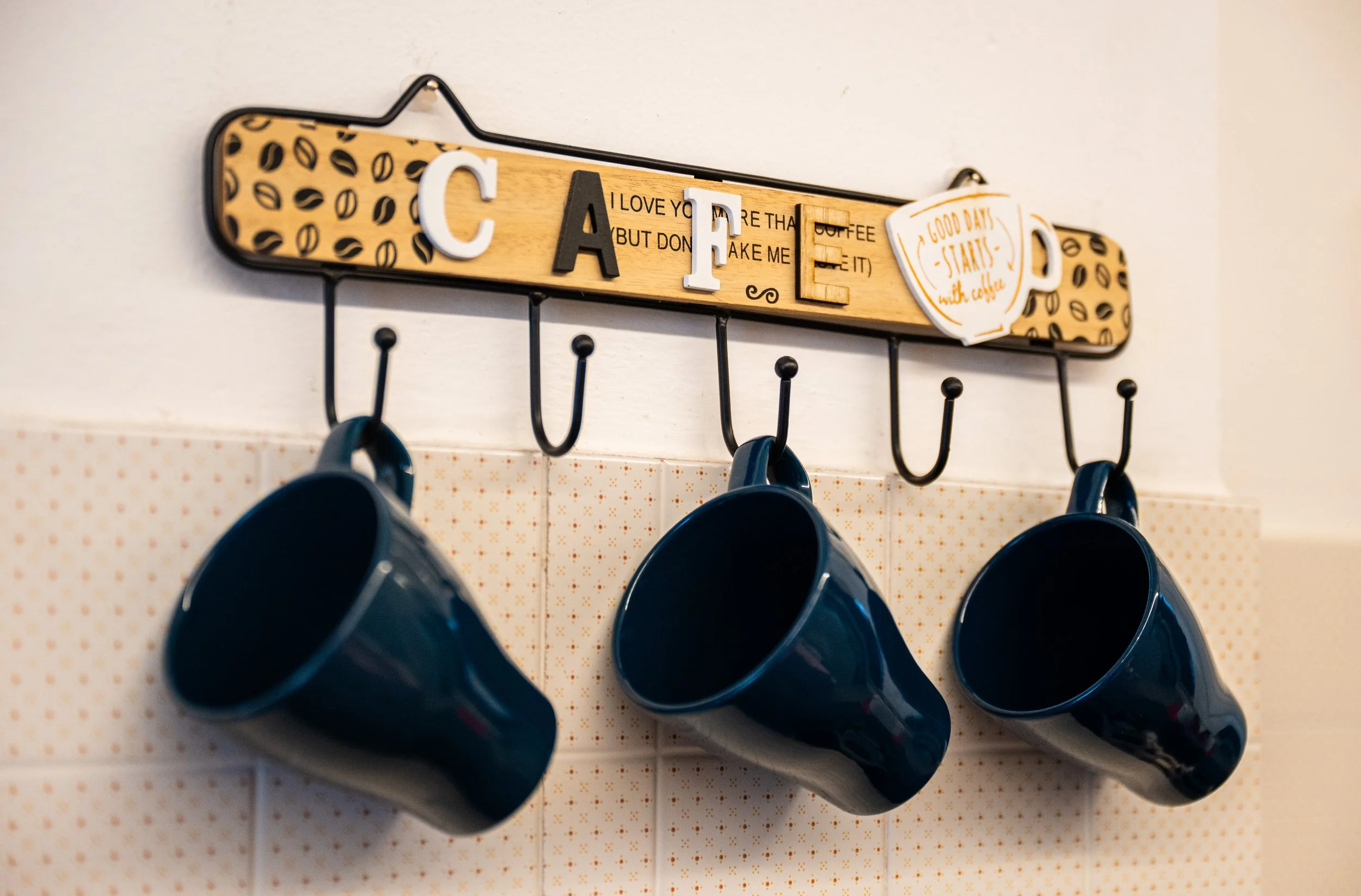Mensola da muro con ganci e tre tazze blu appese, decorazione con lettere e scritto che dice 'Caffè' e 'Good days start with coffee'