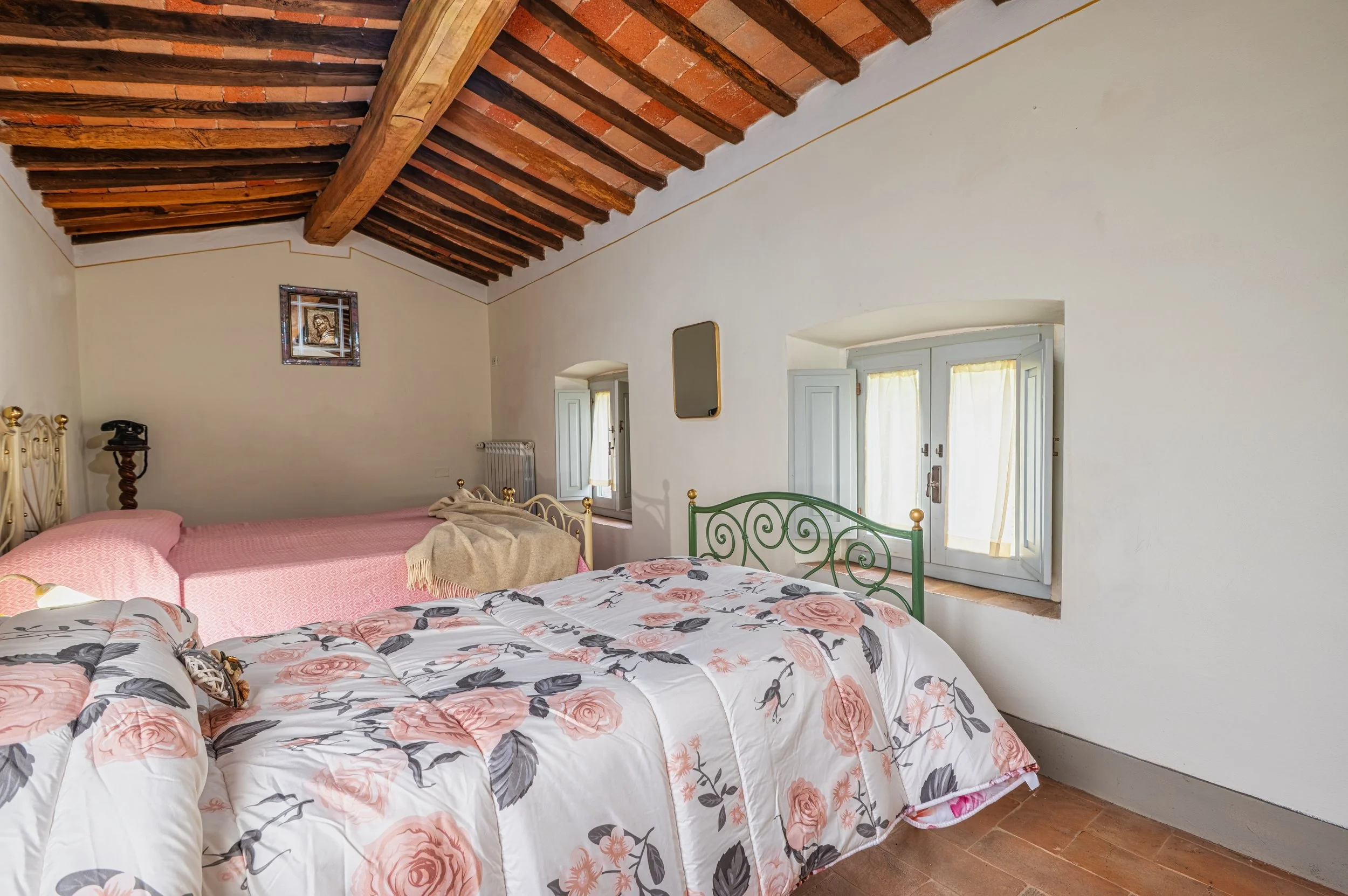 Camera da letto con soffitto in legno, pareti bianche, due finestre con persiane azzurre, letto con copripiumino a fiori rosa e grigi, lampada da tavolo, specchio rettangolare e altri mobili.
