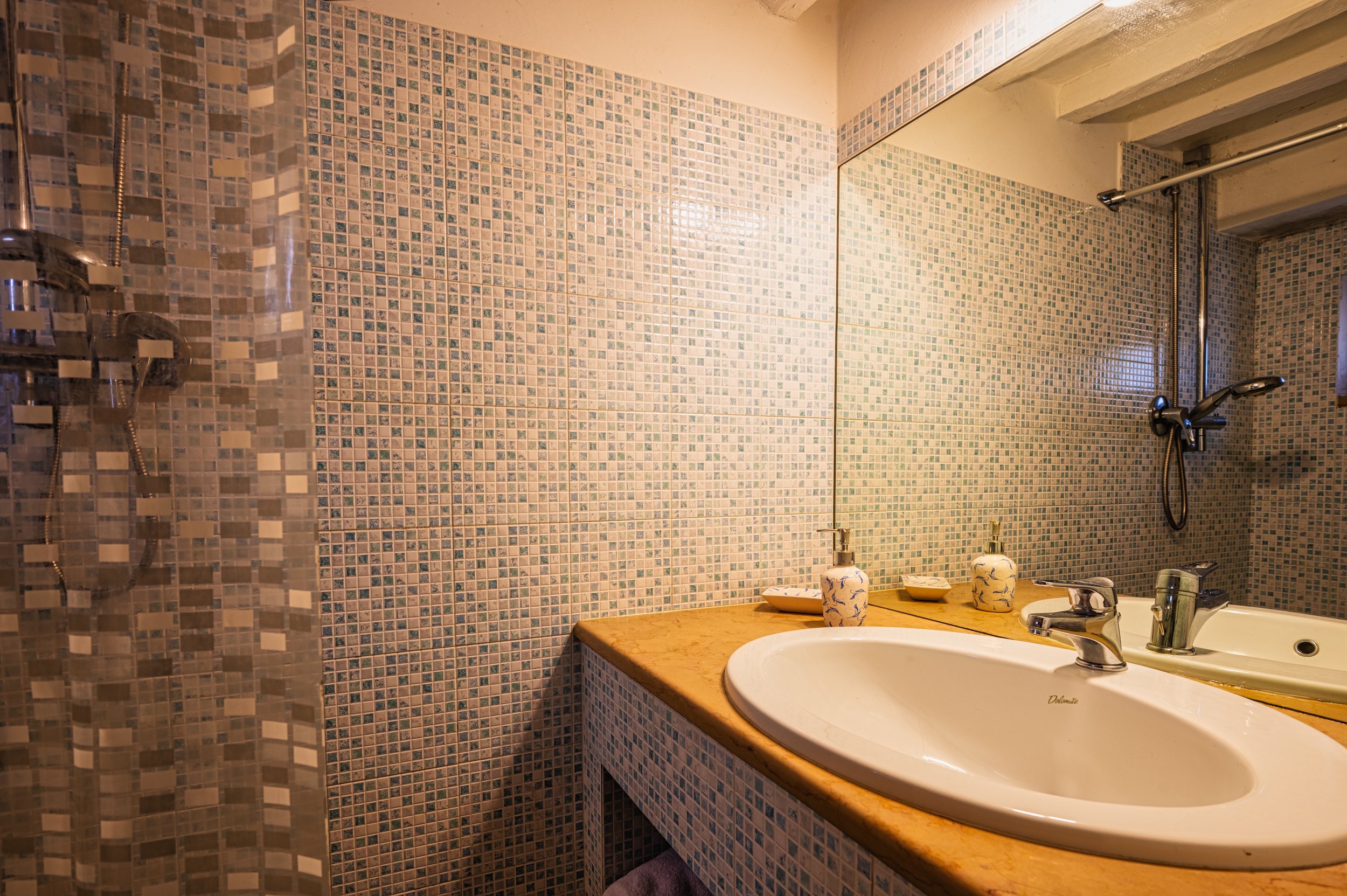Bagno con pareti rivestite di mosaico e lavabo con piani in legno, controsoffitto in cemento, grande specchio e doccia a lato con pareti in mosaico