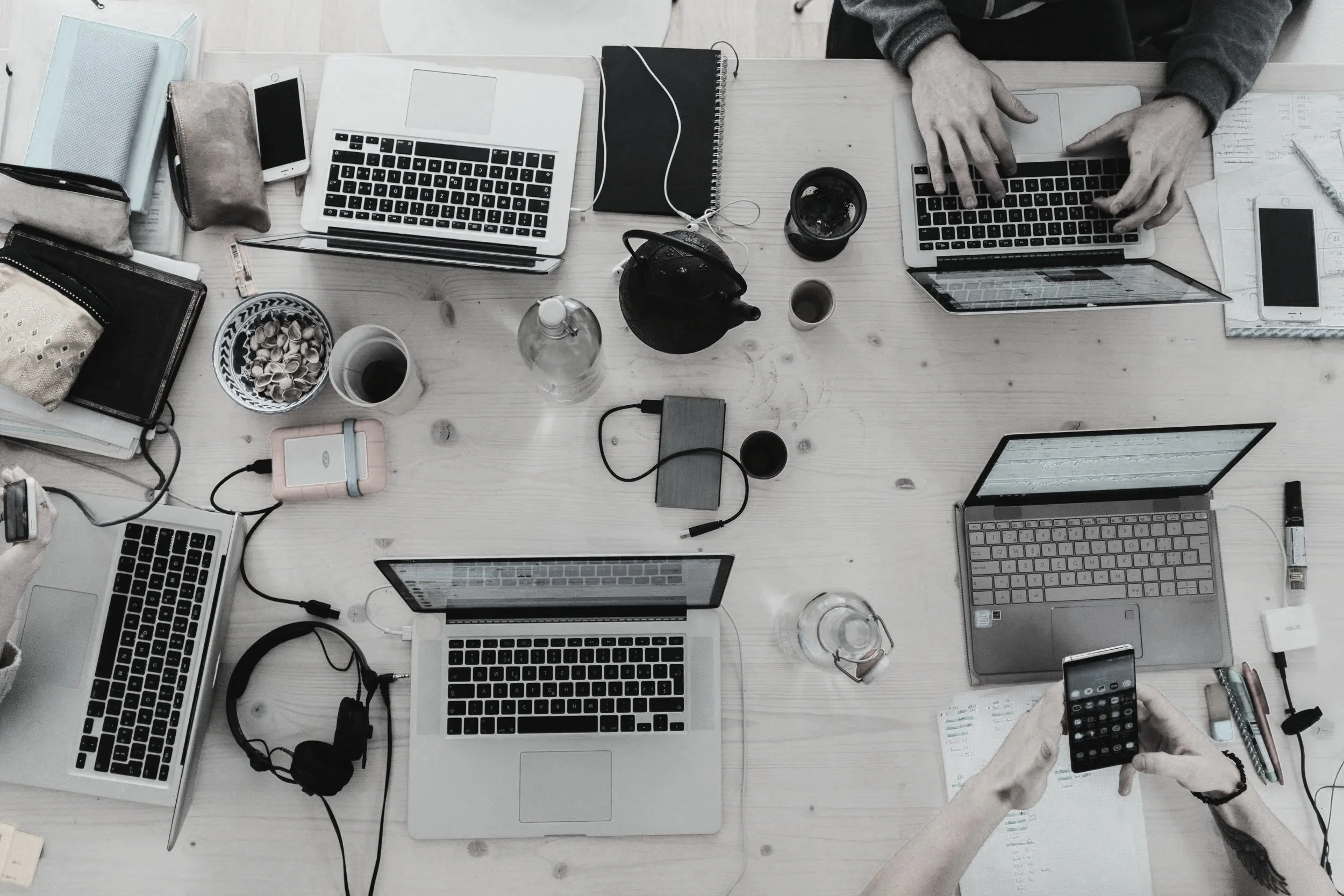Overhead view of a cluttered workspace with multiple laptops, smartphones, notebooks, cups, and various office supplies on a wooden table, with two people working.