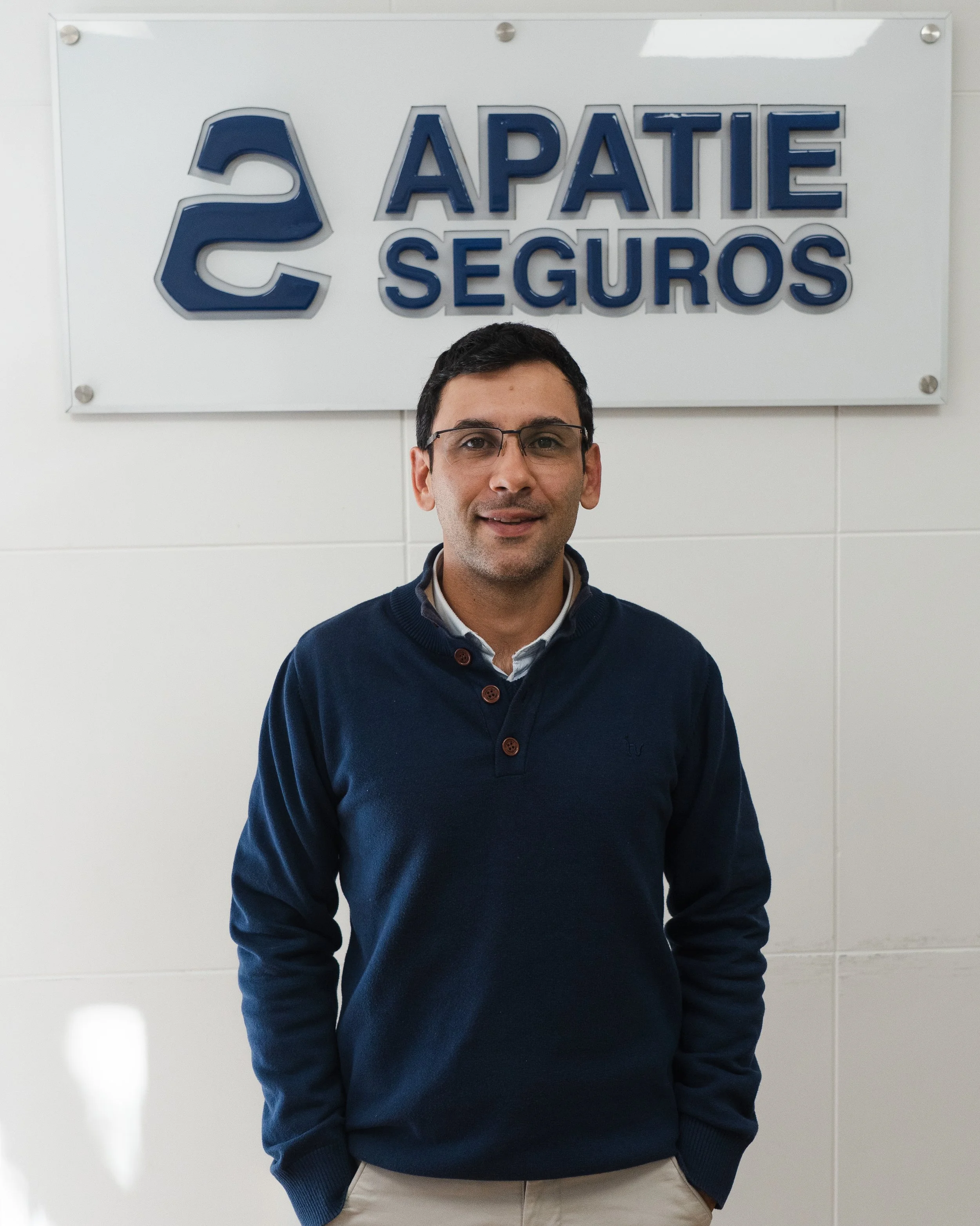 Hombre con gafas, camisa y suéter azul, de pie frente a un cartel que dice 'Apatte Seguros'.