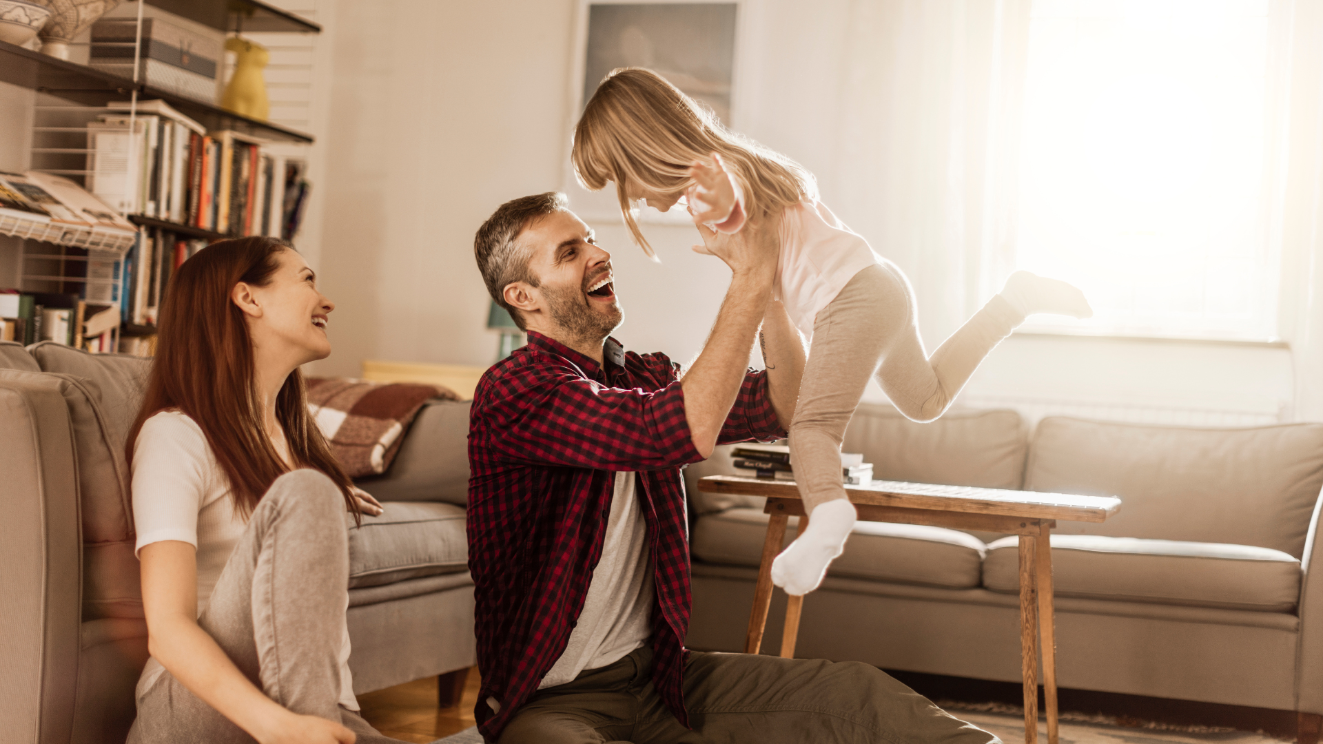 Familia feliz en sala de estar, padre levantando a su hija, madre observando, todos sonriendo.