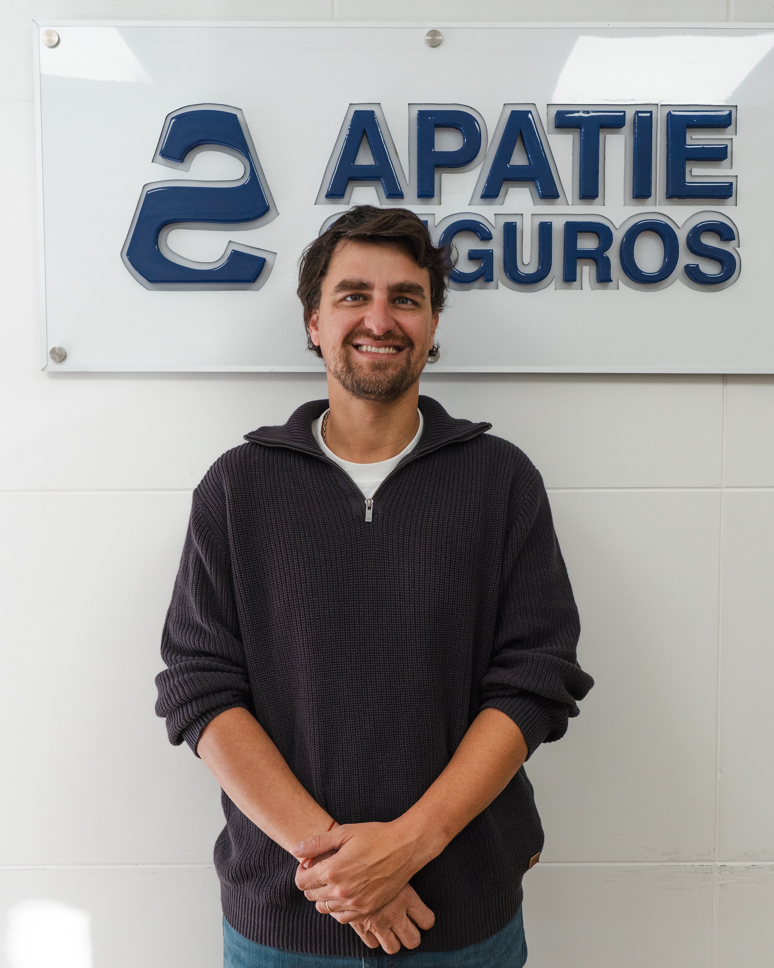 Hombre de cabello castaño oscuro y barba, sonriendo, con una chamarra de punto negra y camiseta blanca debajo, frente a un cartel con letras azules que dice 'S APATIE SEGUROS'.