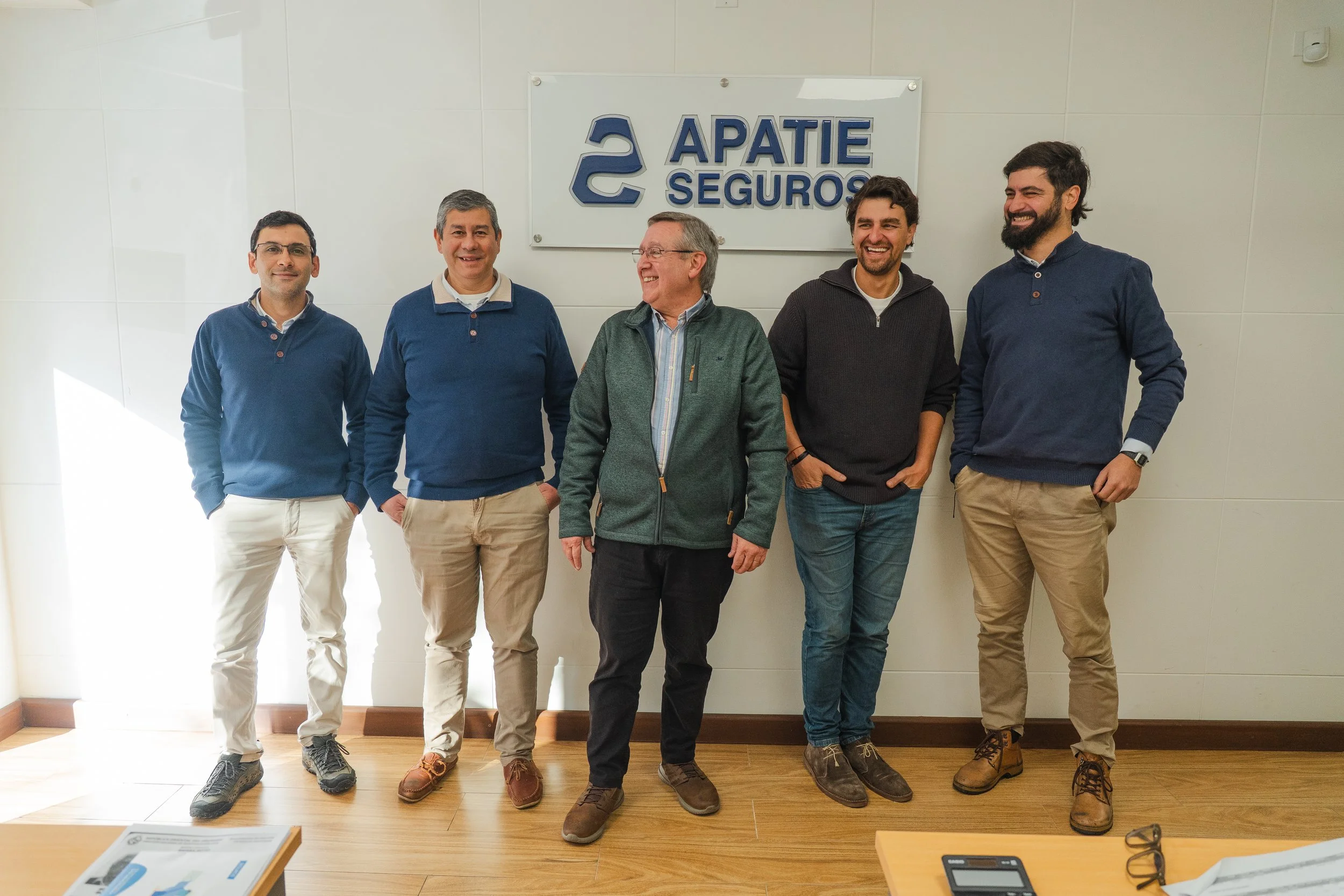 Cinco hombres de pie frente a un cartel que dice 'APATIE SEGUROS', en una oficina, relajados y sonriendo, con vestimenta casual y fondo blanco.