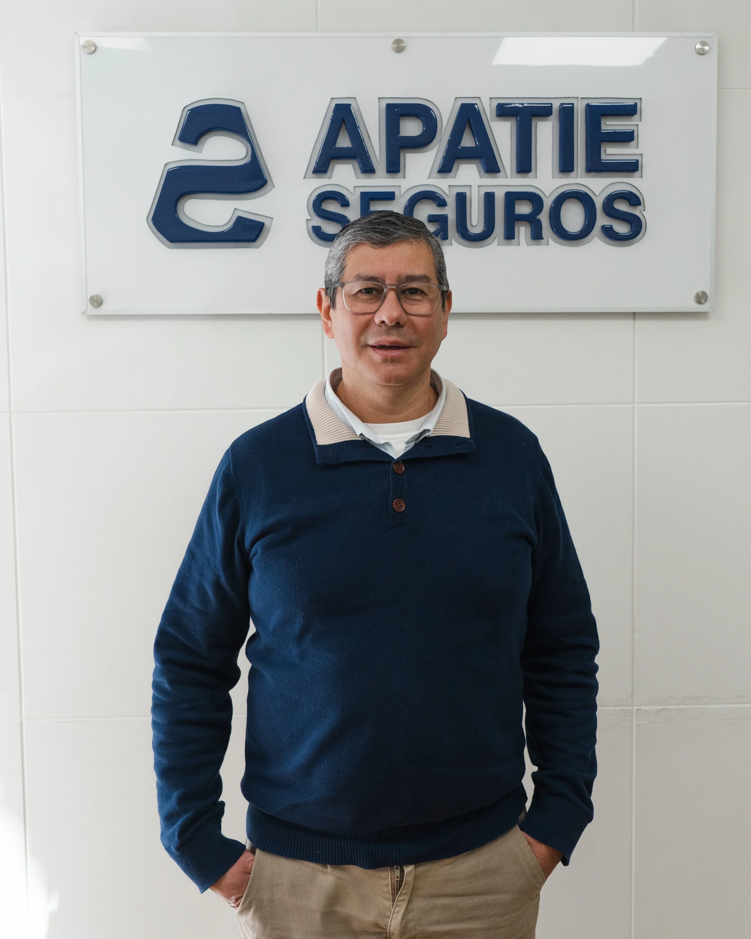 Hombre de pie frente a un cartel que dice 'APATIE SEGUROS' con un logo azul, en una pared blanca, con manos en los bolsillos, usando gafas, suéter azul y camisa blanca.