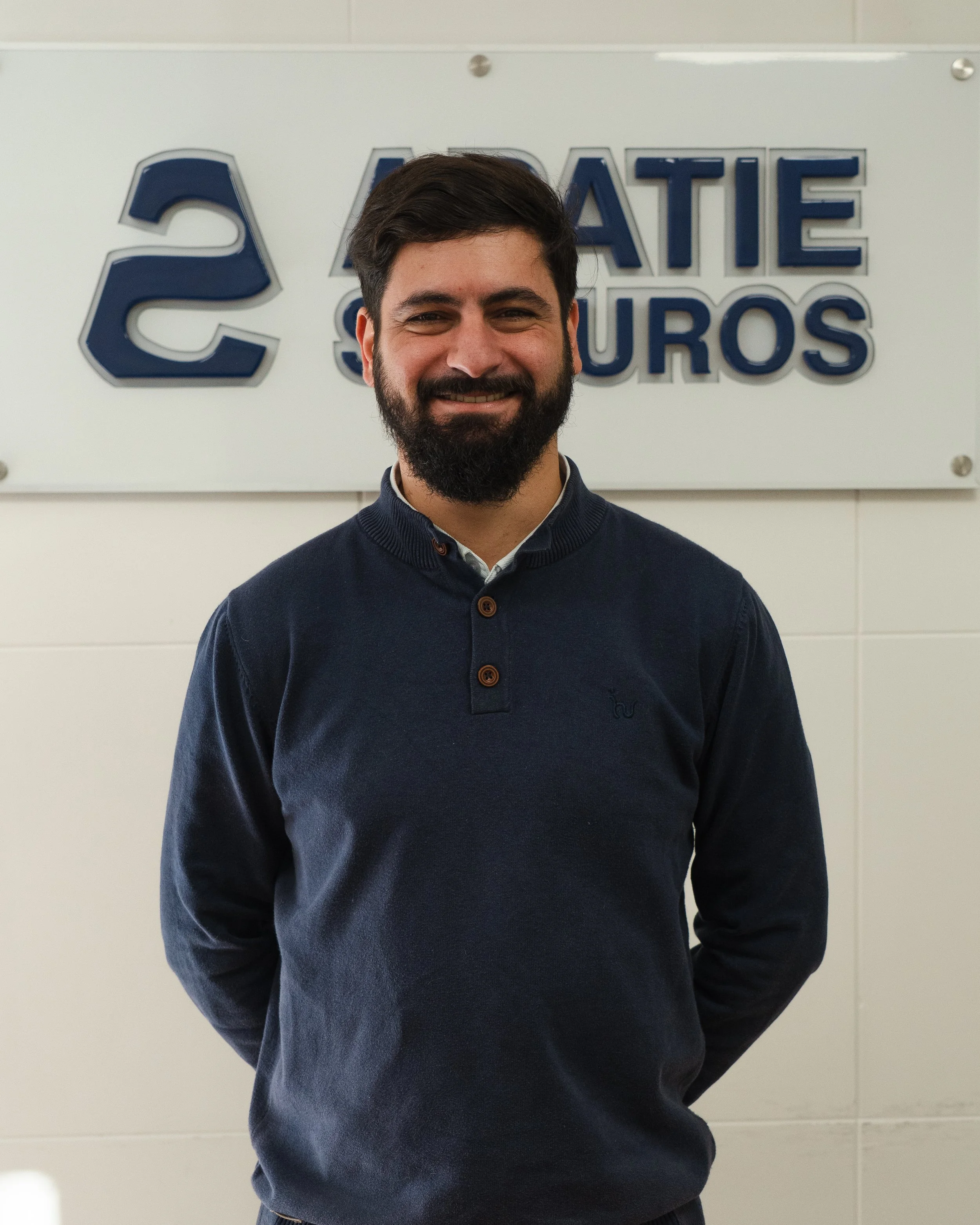 Un hombre de cabello oscuro y barba, con una camisa blanca y suéter azul, de pie frente a una pared con letrero que dice 'Ahorate 2 Seguros'.