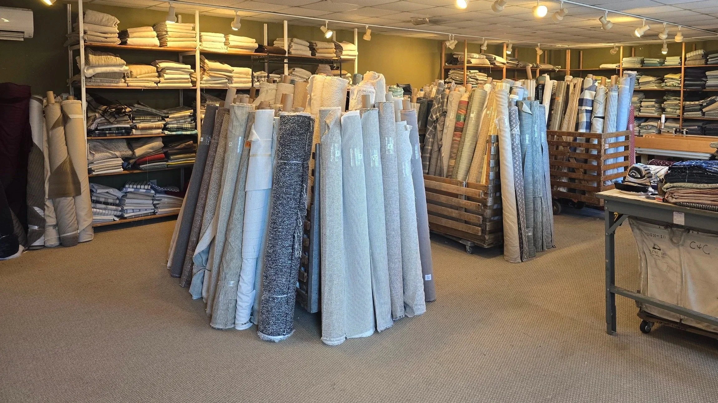 Interior of a fabric store with shelves and racks filled with various bolts of fabric, including rolled and folded pieces, in a range of colors and patterns.