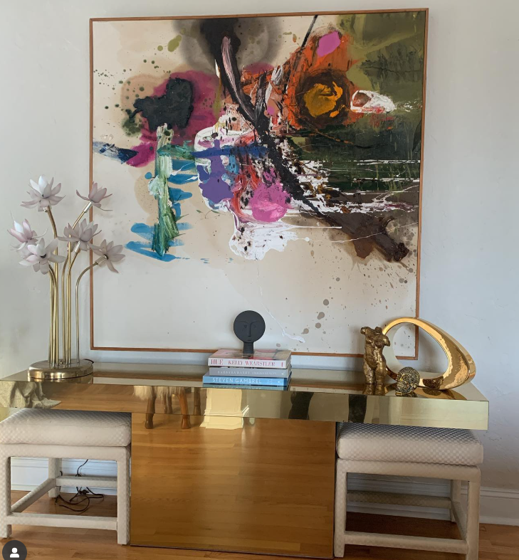 Contemporary living room decor featuring a large abstract painting with vibrant colors on the wall, a gold and white sideboard with decorative items including books, sculpture, and accessories, and a flower sculpture with pink and white blooms on a stand.