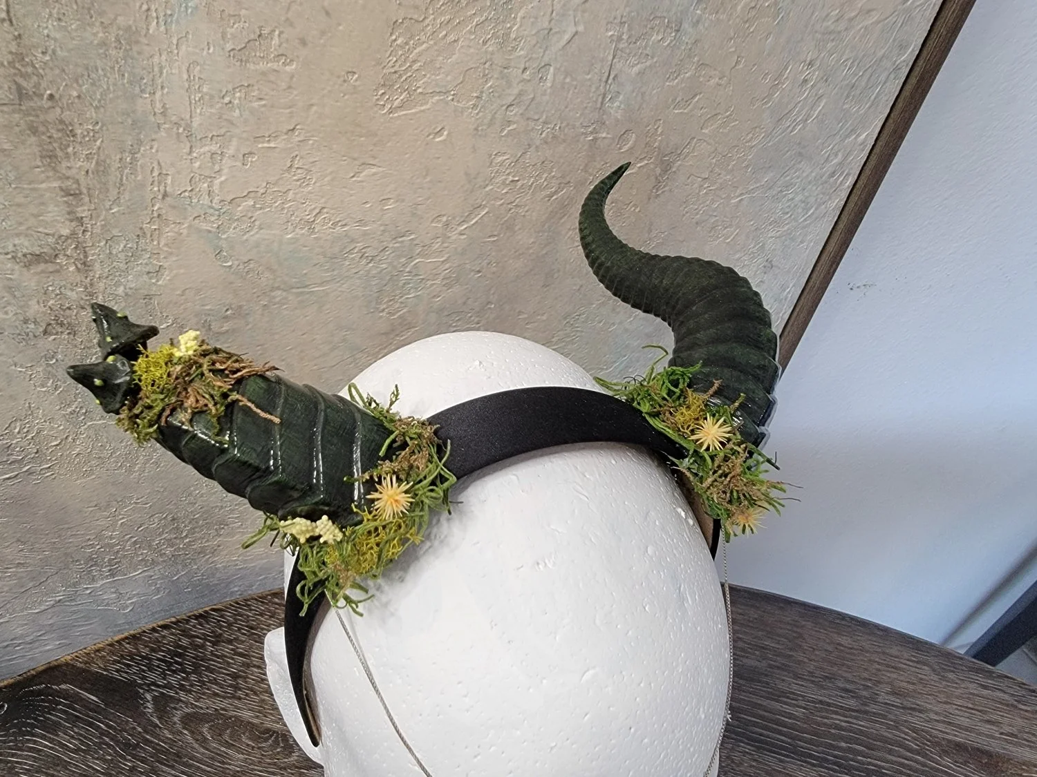 Larger dragon-style horns, that are black/dark-green, mounted on a black headband. One horn is broken and the top of the broken horn appears to be covered in moss and growing mushrooms. The base of the horns are also surrounded by moss, with yellow f