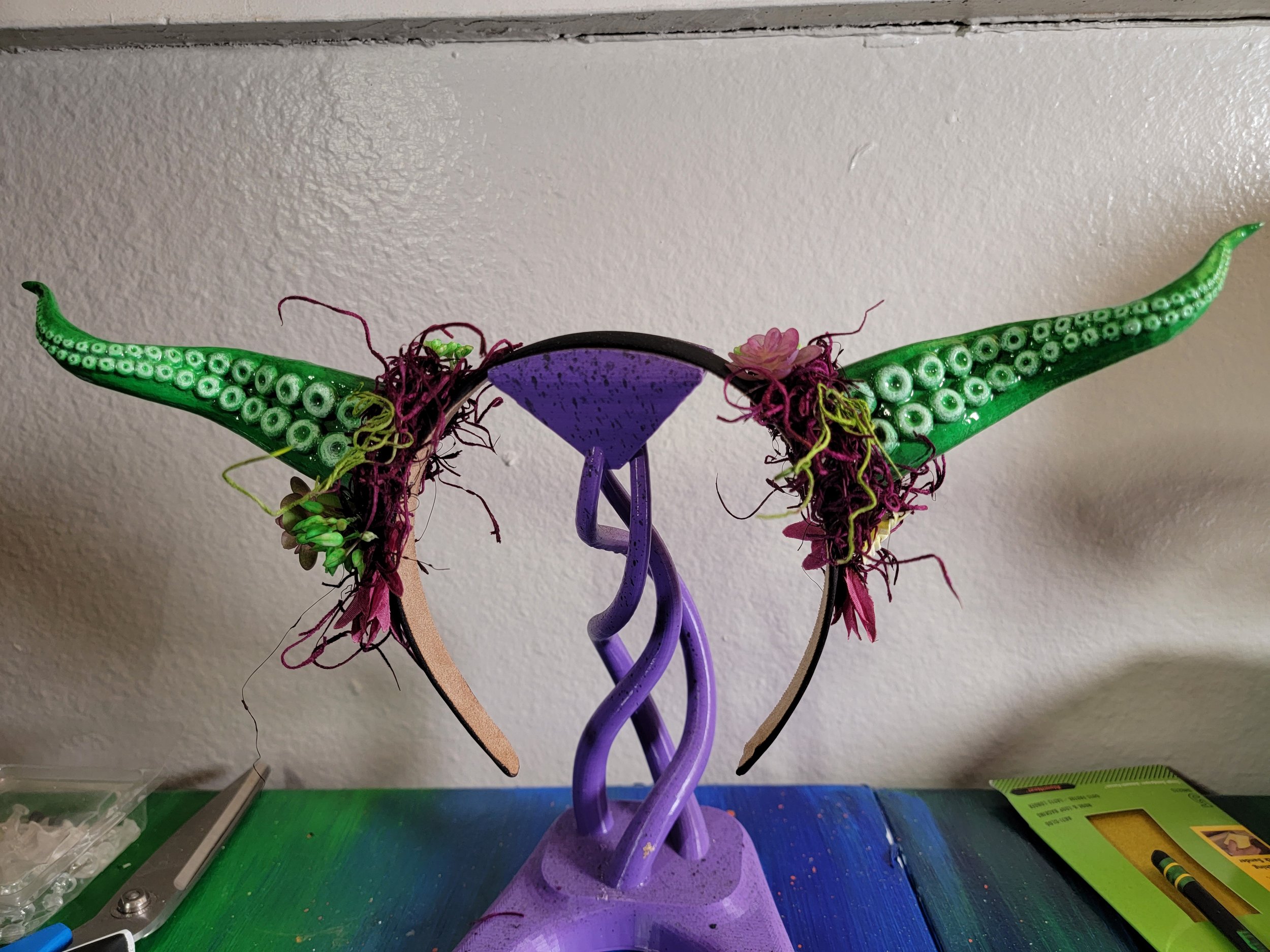 Green tentacle horns, mounted on a headband, and each tentacle horn's base is covered in a dark purple/magenta moss, with floral elements.
