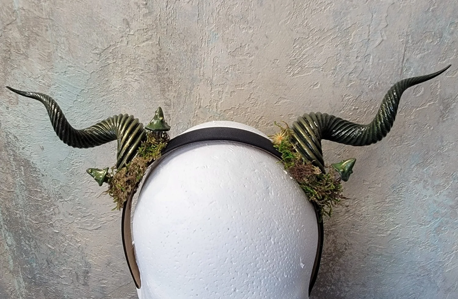 Metallic green spiral horns on a Styrofoam mannequin head. Each horn's base is surrounded by moss that appears to be growing around it, with mushrooms and tiny yellow flowers appearing to grow out of the moss.
