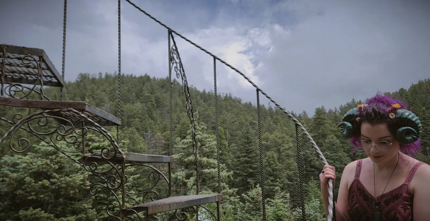 a photograph of a woman wearing dark teal ram's horns holding onto a spiral staircase with a coniferous forest behind her