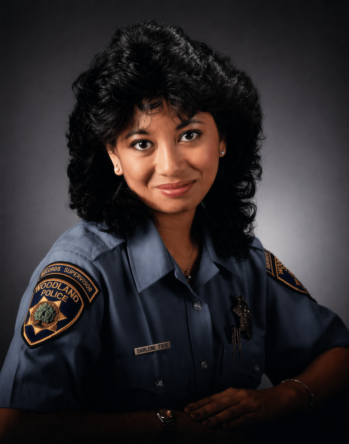 Portrait of a female police officer with black curly hair, wearing a dark blue uniform with patches and a badge, against a gray background.