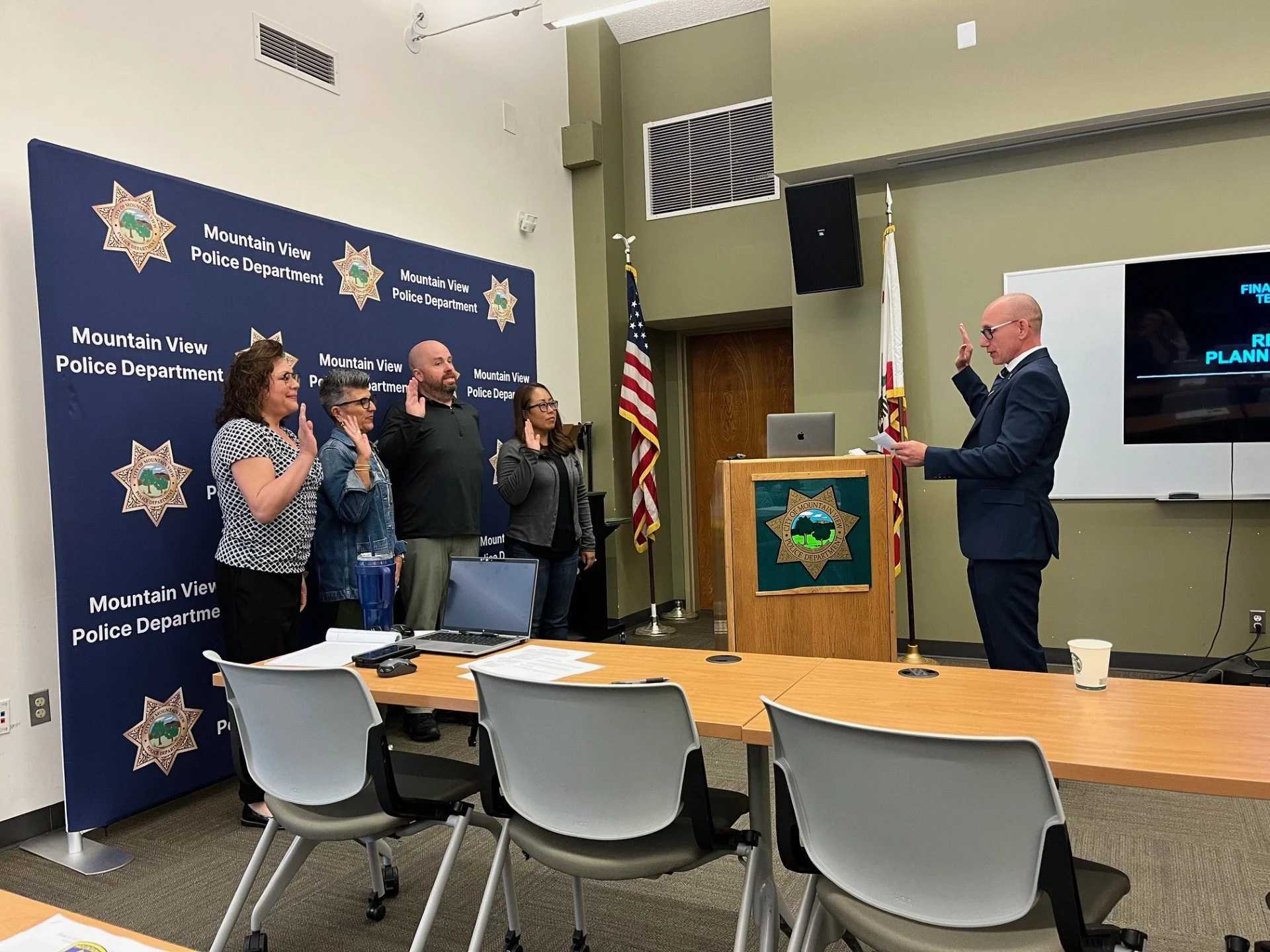 A group of five people standing before a blue backdrop with the logo and text 'Mountain View Police Department'. A man in a suit is raising his right hand, reading from a paper, while the others, three women and one man, are taking an oath with right hands raised. An American flag and a California state flag are visible behind them. The room has beige walls, visible chairs, and a table with papers and a laptop.