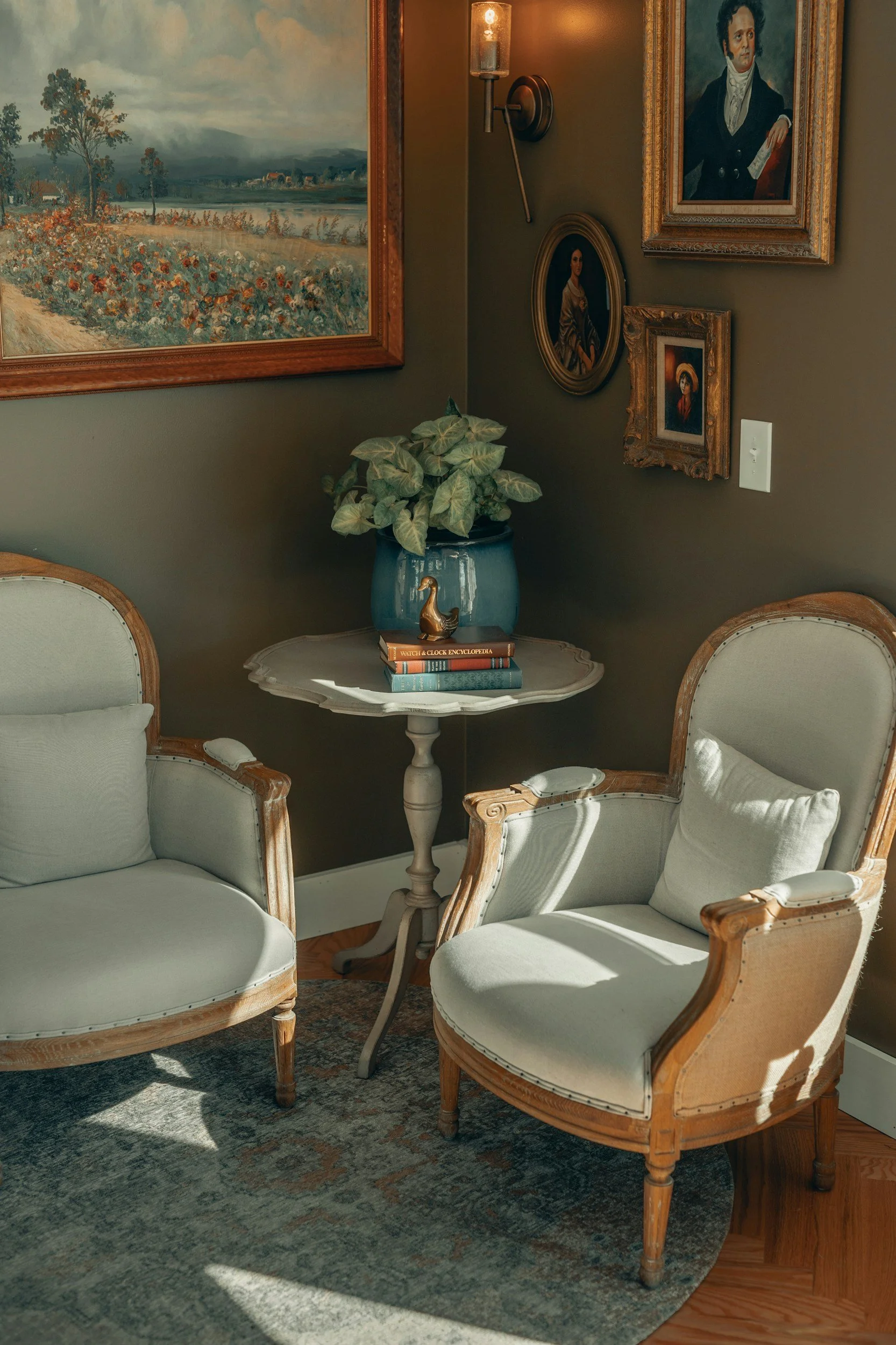 A cozy sitting area with two vintage armchairs with white upholstery and wooden frames, a small round white table with books and a potted plant, and framed artwork on a brown wall illuminated by a wall sconce.