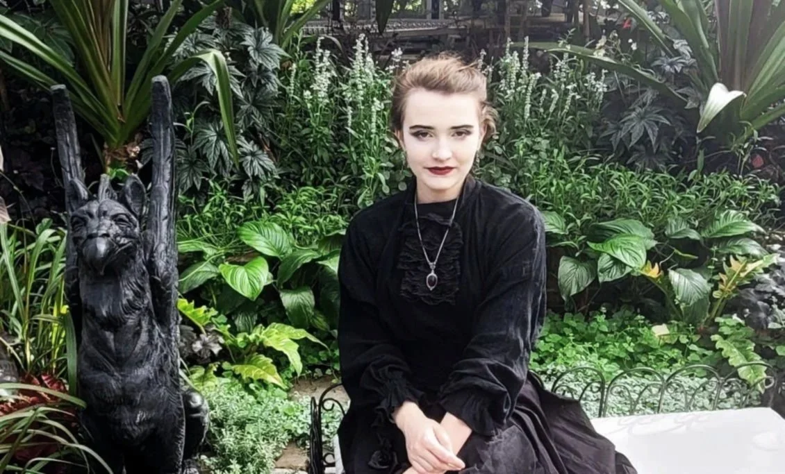 A young woman with brown hair, wearing a black dress and necklace, sitting outdoors among lush green plants, with a black statue of a dog with one paw raised to her left.