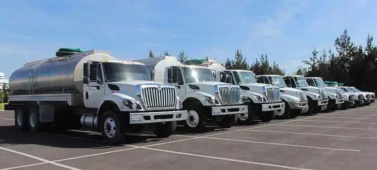 A row of white fuel or water delivery trucks parked in a parking lot under a blue sky.