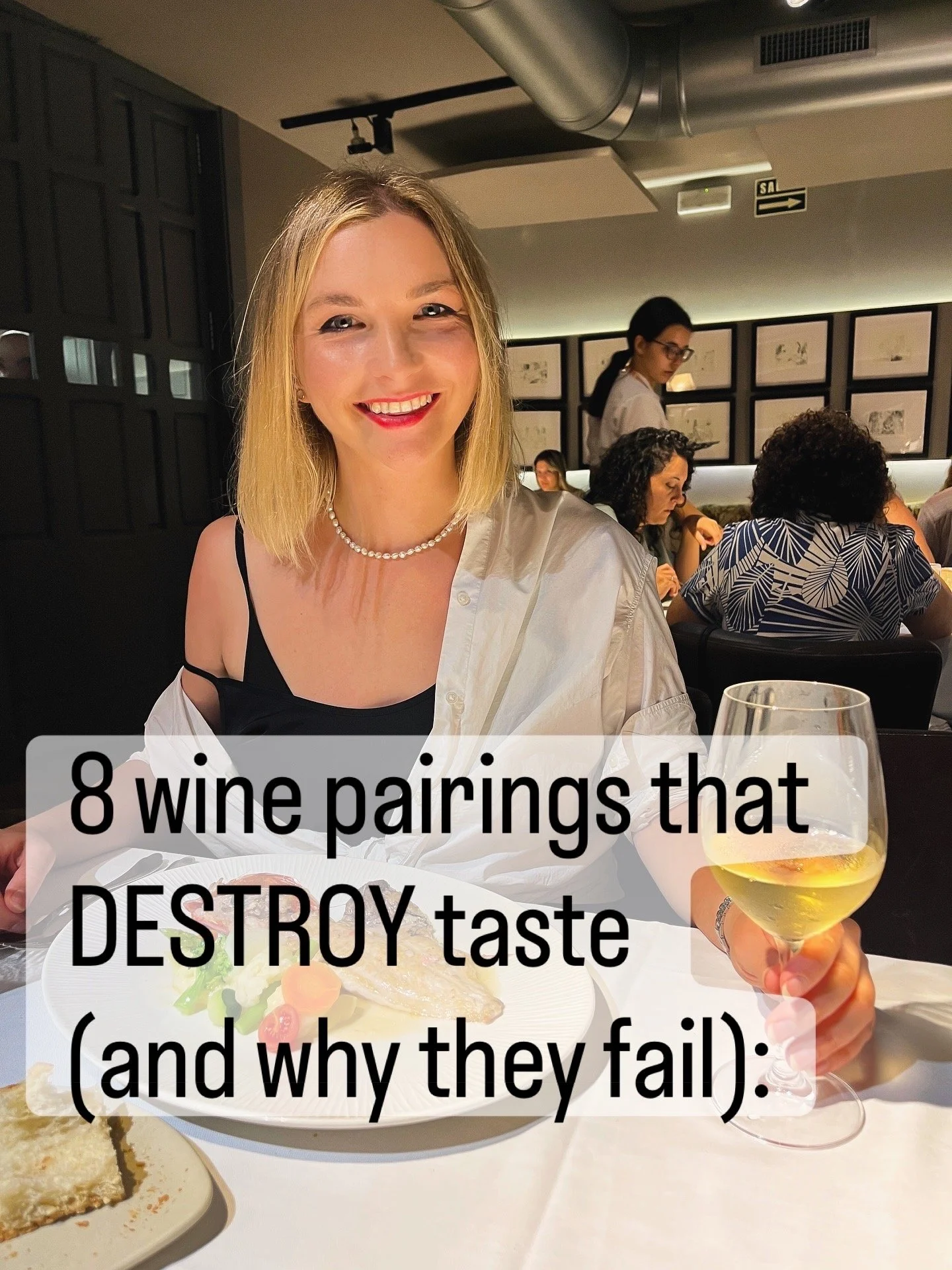 A woman with blonde hair smiling at a dining table, holding a glass of white wine, with a plate of food in front of her, in a restaurant setting with other diners in the background.