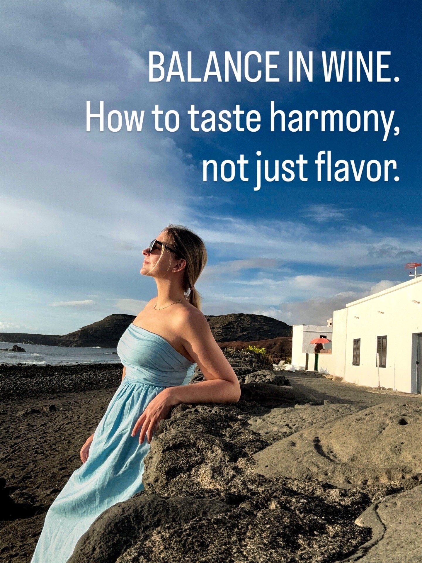 A woman in a light blue strapless dress and sunglasses leaning on rocks at the beach with white buildings, a windmill, and a hill in the background under a partly cloudy sky. Text overlay reads: "Balance in wine. How to taste harmony, not just flavor."