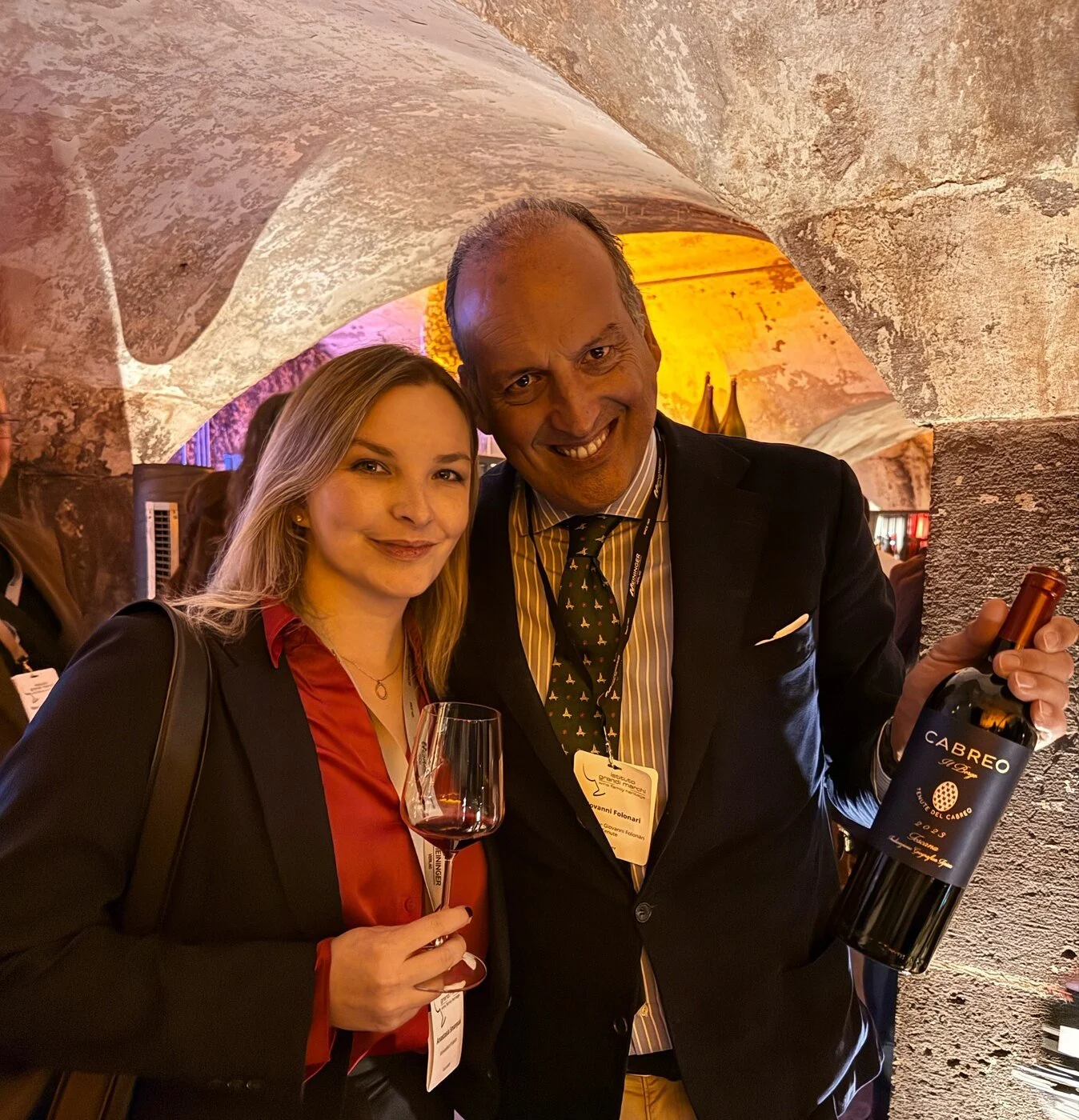 Giovanni Folonari of Ambrogio e Giovanni Folonari Tenute holding a bottle of Cabreo Il Borgo 2023 Toscana IGT, smiling next to a woman with a glass of red wine in the stone cellar of wineBANK Köln
