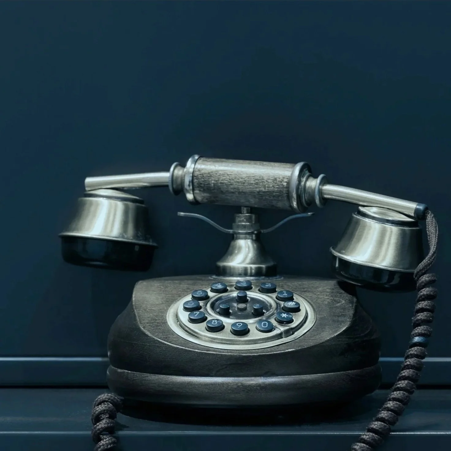 Vintage rotary phone with a dark wooden base, metallic body, and curled cord, set against a dark background.