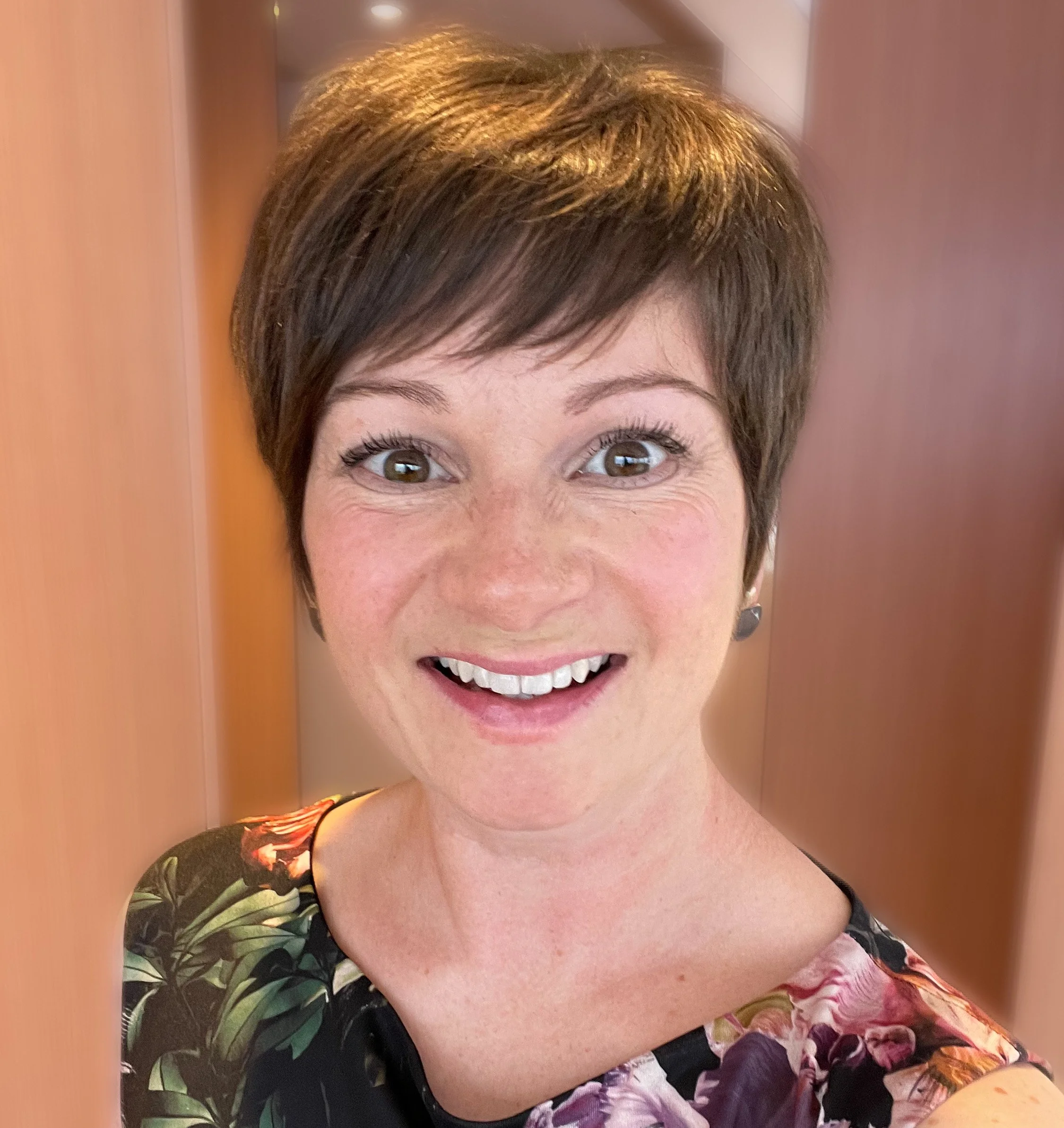A woman with short brown hair, smiling, wearing a floral top, standing between wooden panels.