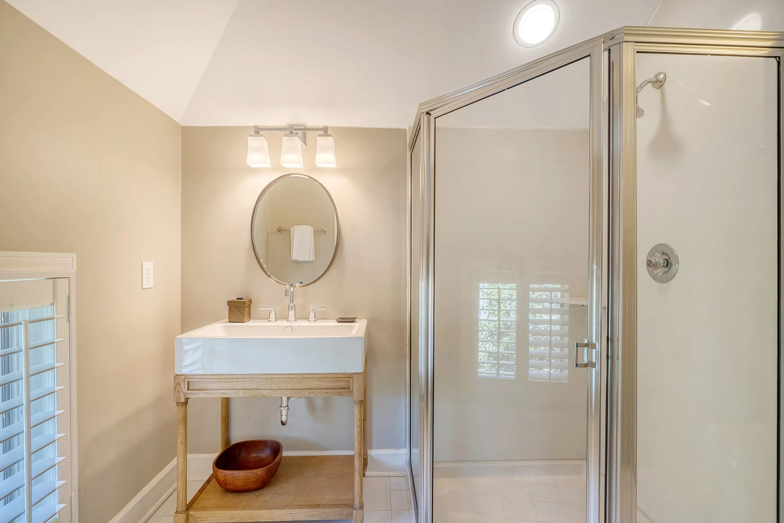 Bathroom with beige walls, a small wooden vanity with a white sink, a round mirror above the sink, a light fixture with three bulbs, a wicker container on the vanity, a brown wooden bowl underneath, a shower with a glass door to the right, a window w