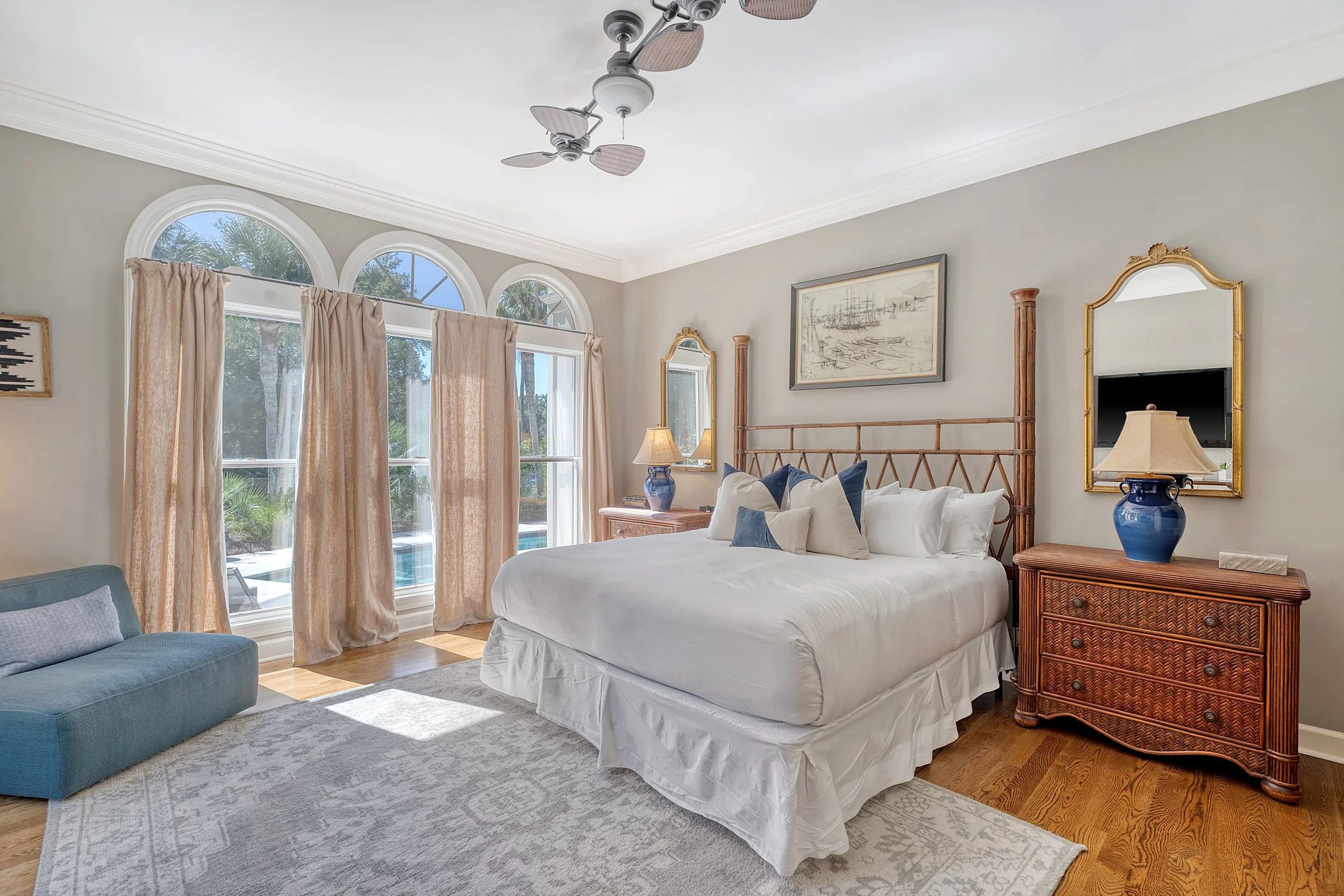 A bedroom with a large bed, blue and white pillows, wooden side tables with lamps, window with beige curtains, and tropical greenery outside.
