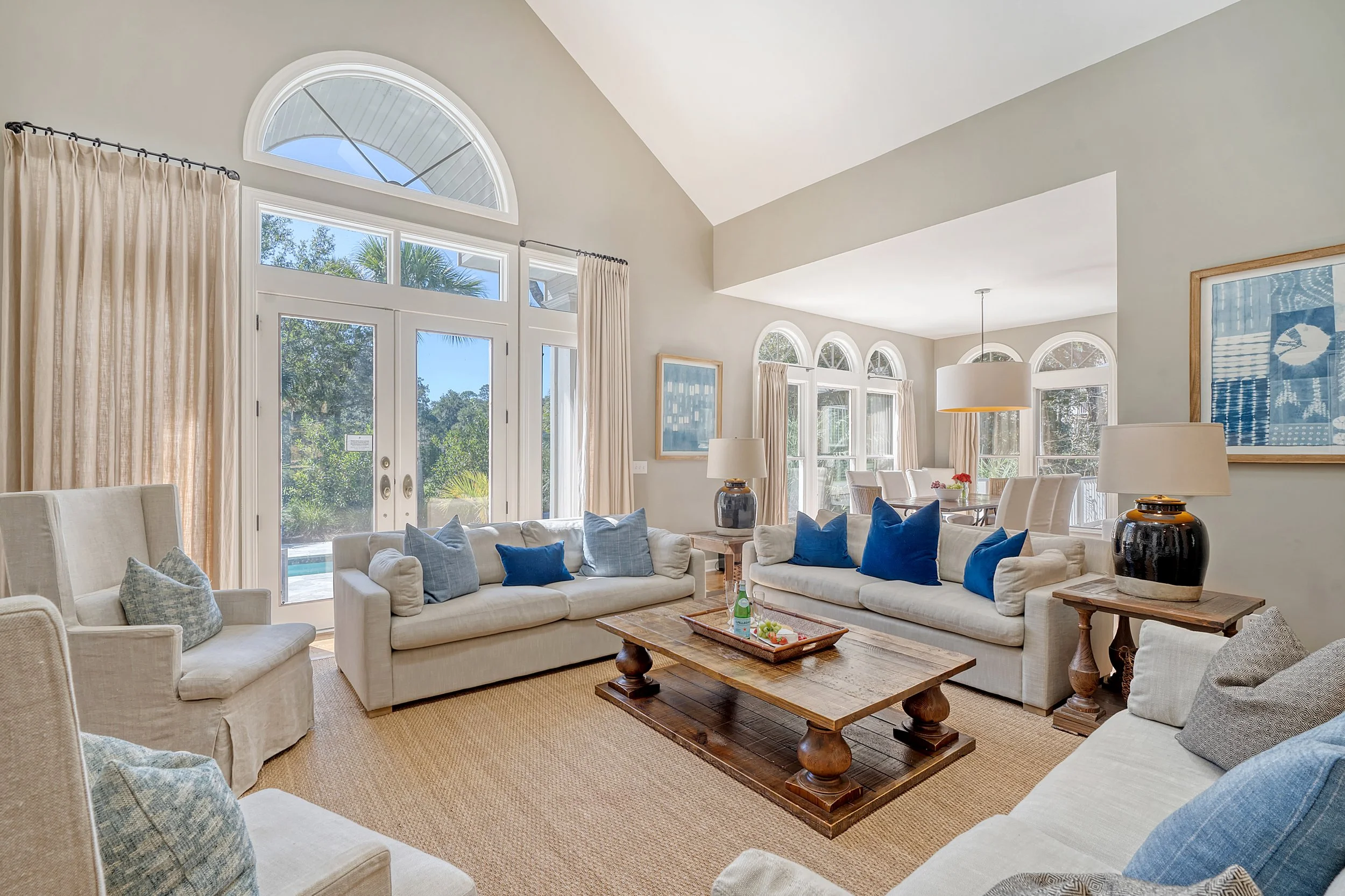 Bright living room with large windows and patio door, beige sofas with blue and gray cushions, wooden coffee table, side tables with lamps, and dining area in the background.