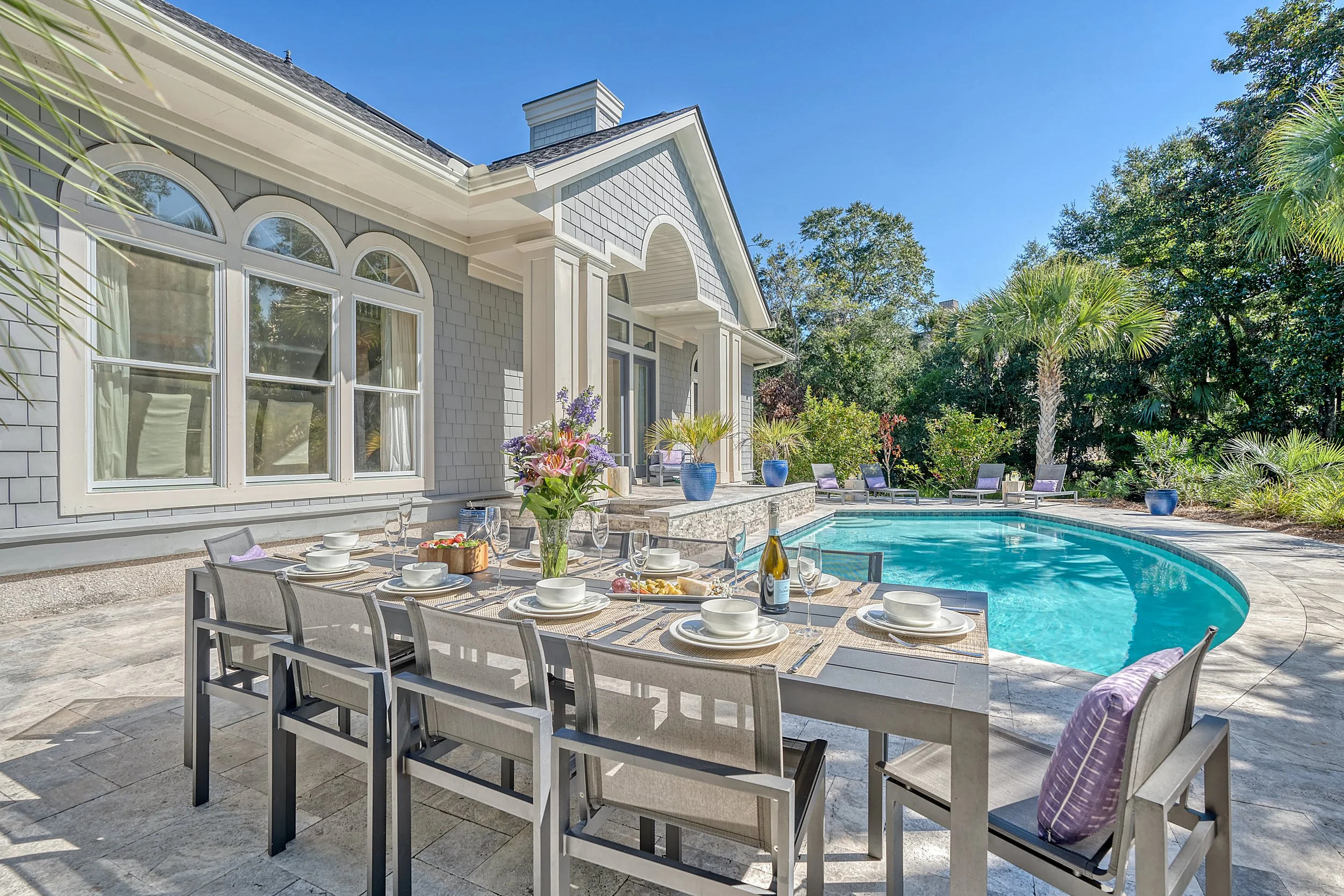 An outdoor patio with a dining table set for a meal, including plates, glasses, and a bottle of wine. The patio features a large swimming pool surrounded by lounge chairs and lush greenery, with a large house in the background.
