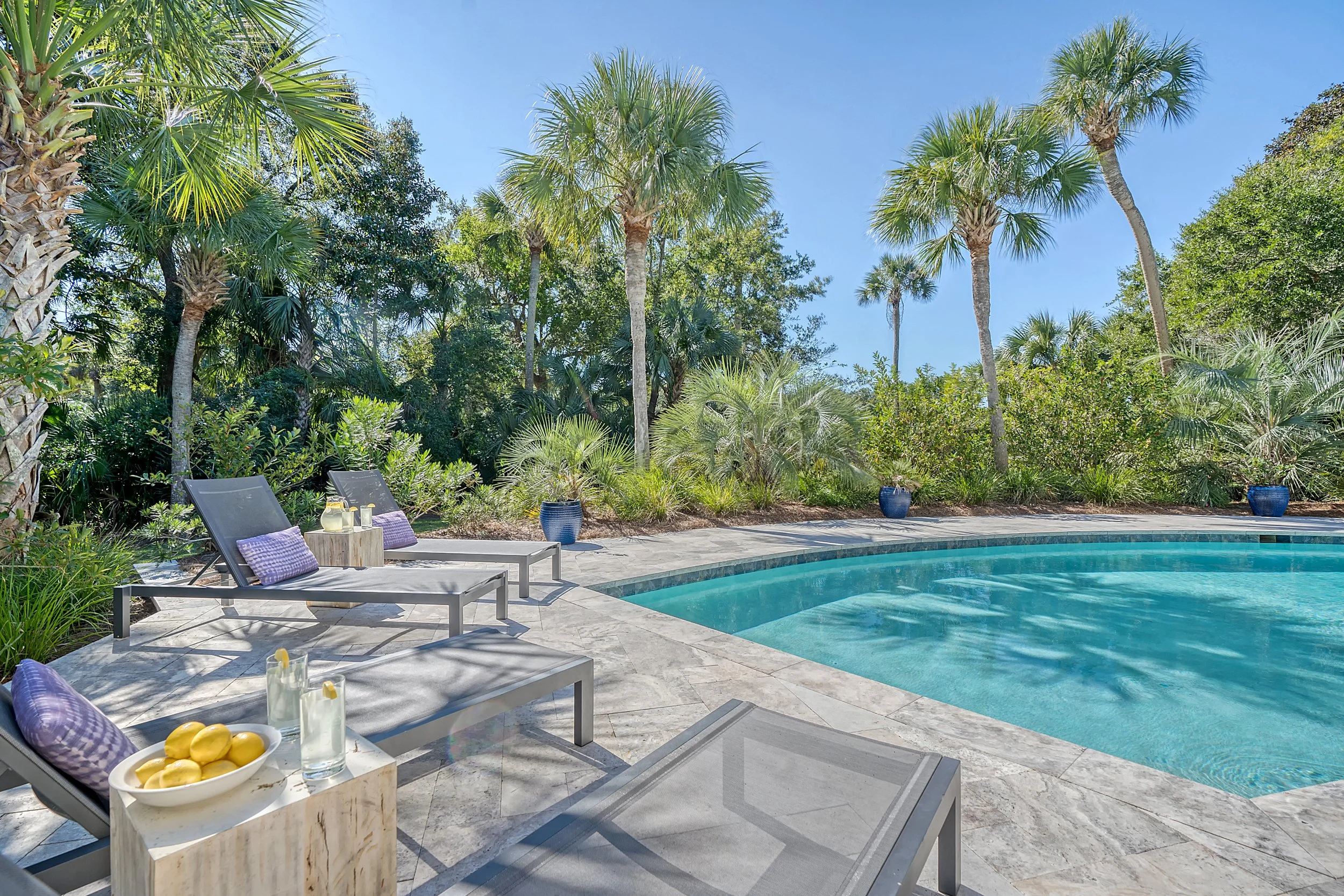 Poolside area with lounge chairs, purple pillows, and small tables with drinks and lemons, surrounded by tropical plants and palm trees under a clear blue sky.