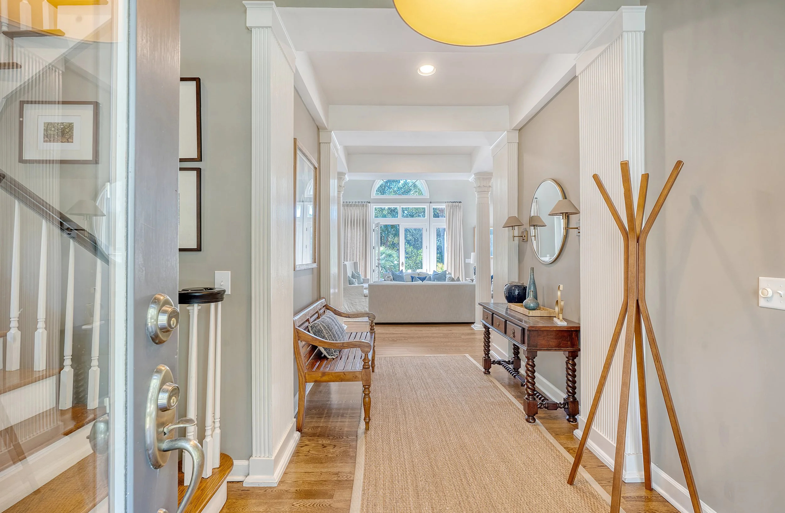 View of a bright and elegant entryway leading into a living room with large windows and white curtains, wooden furniture, and decorative vases.
