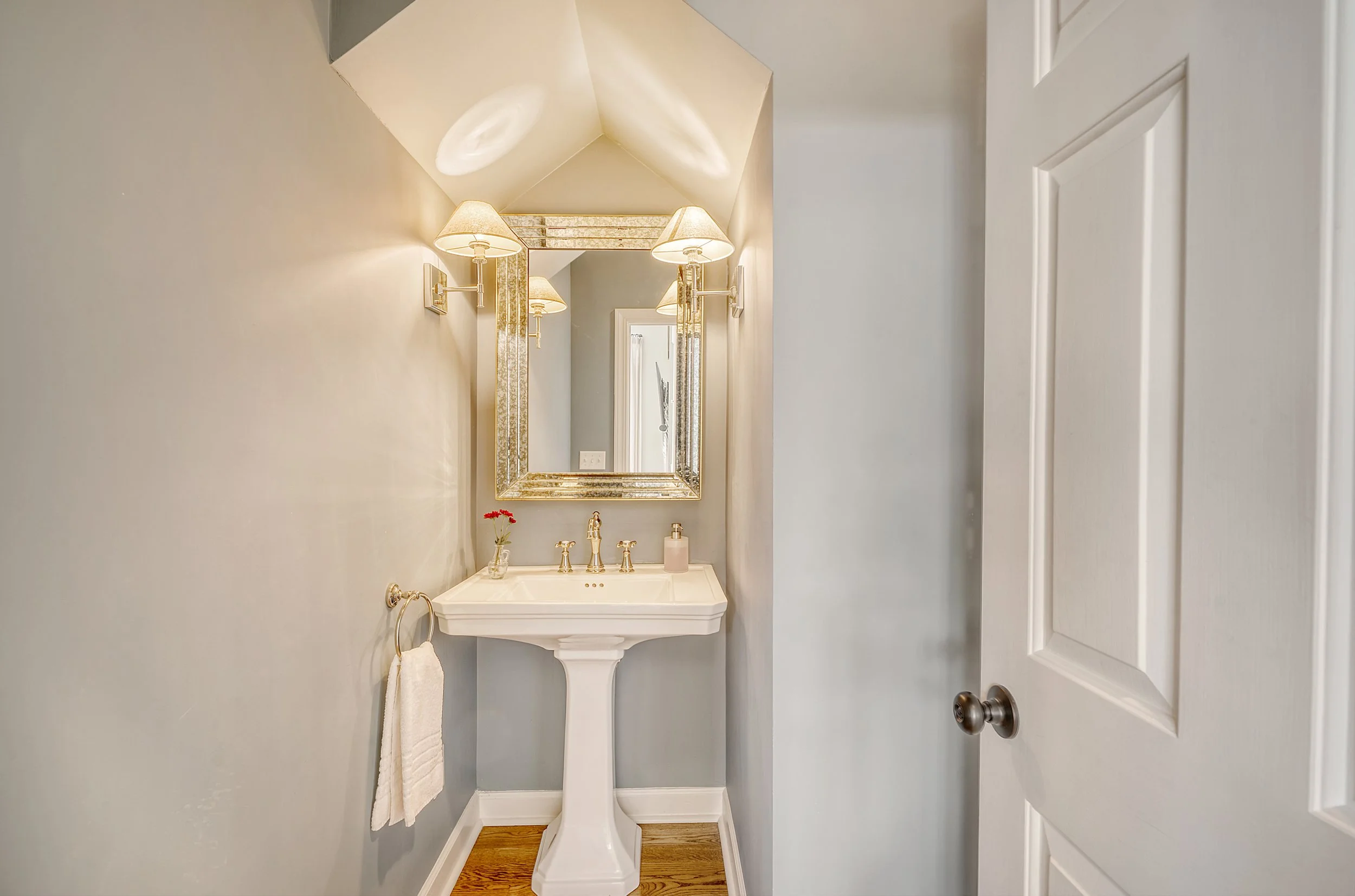 Small bathroom with a pedestal sink, gold fixtures, a large mirror, two wall sconces, and light-colored walls.