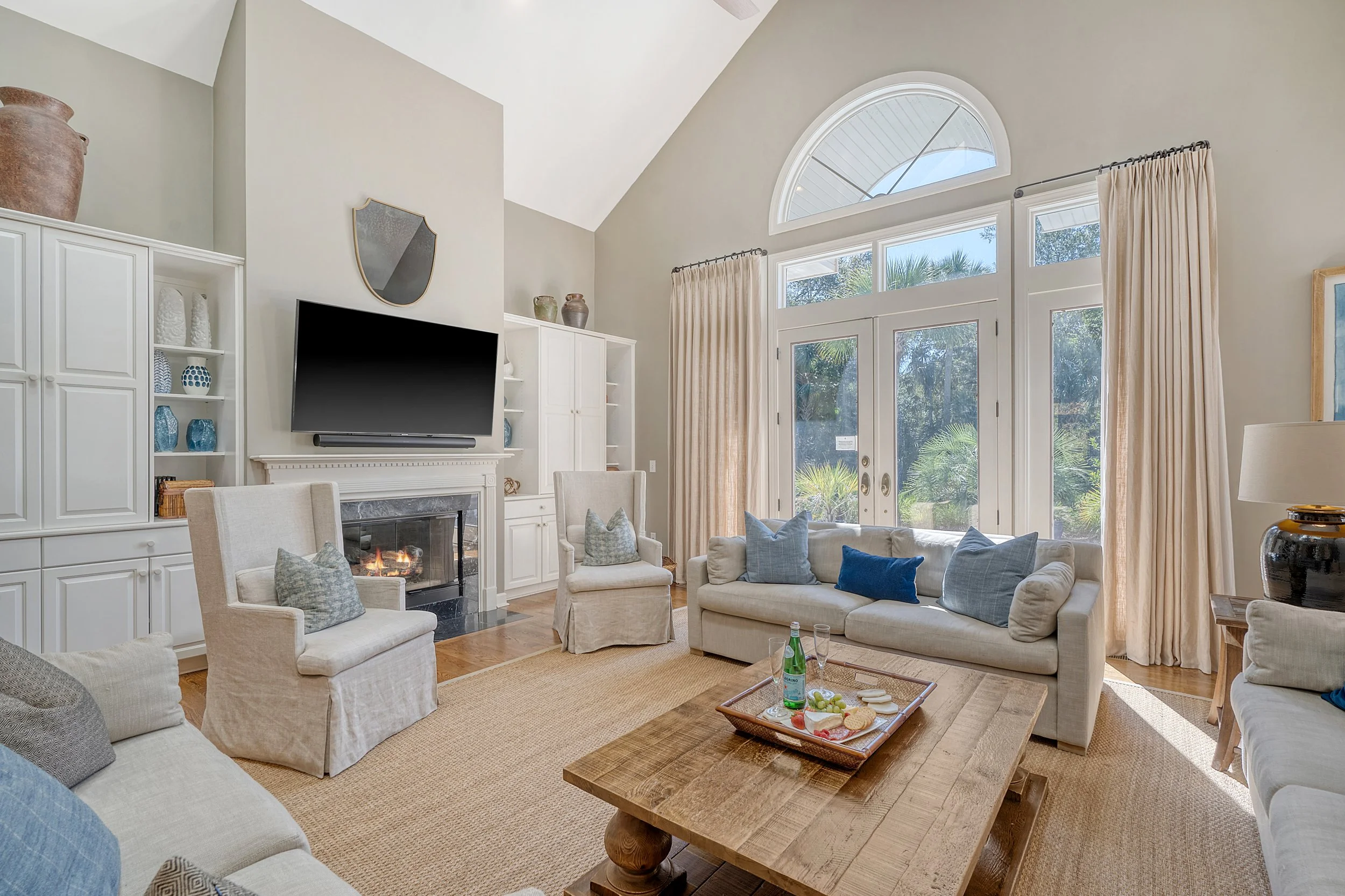 Living room with white sofas and chairs, a wooden coffee table with snacks and drinks, a fireplace, a large flat-screen TV, large windows with beige curtains, and a neutral color scheme.