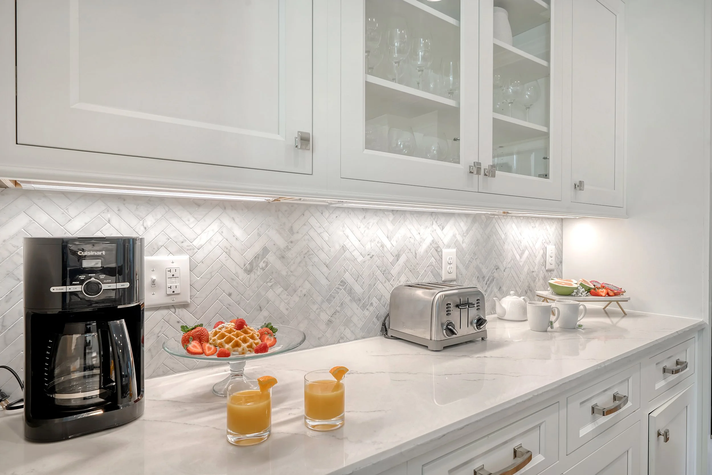 White kitchen countertop with a coffee maker, two glasses of orange juice, a fruit plate with waffles and strawberries, a toaster, a teapot, two mugs, and a tray of strawberries and other fruits, with white cabinets and a marble backsplash.