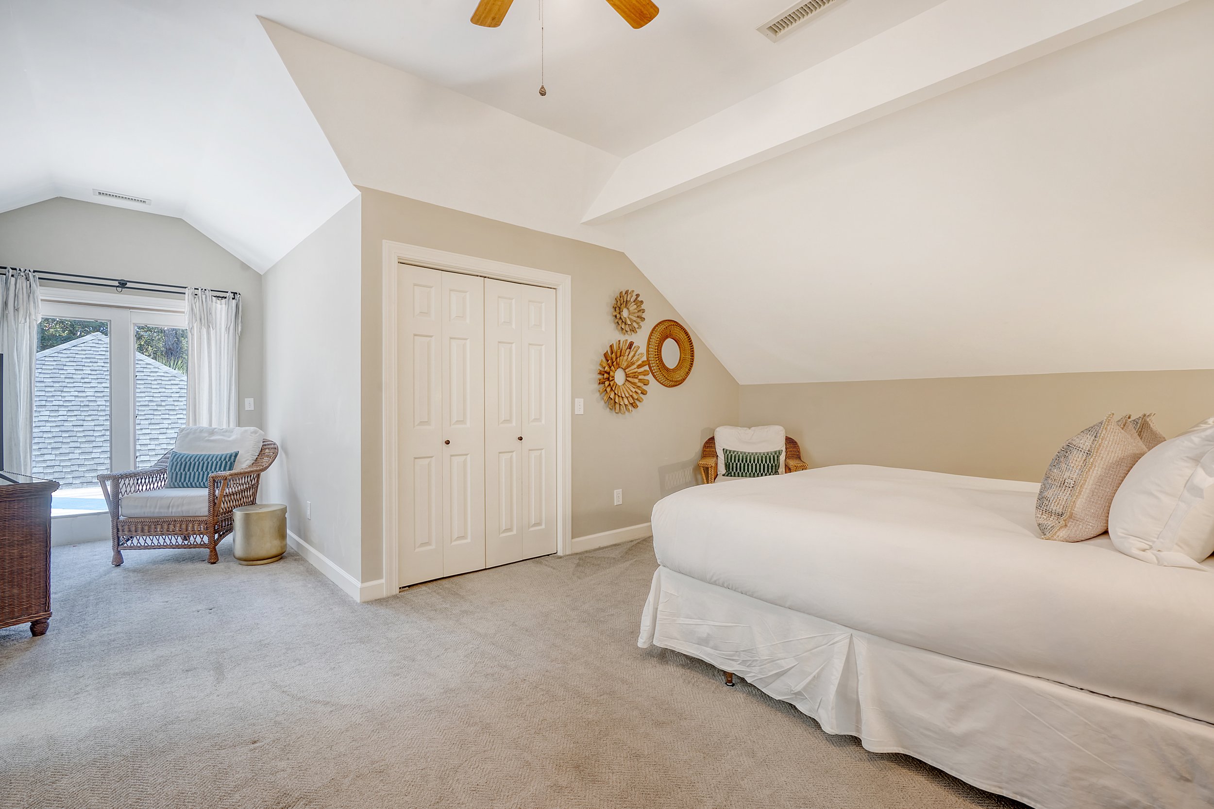 A bedroom with a sloped ceiling, a bed with white linens and pillows, a small sitting area with wicker chairs and decorative items on the wall, beige carpet, and a sliding glass door with curtains leading to an outside view.