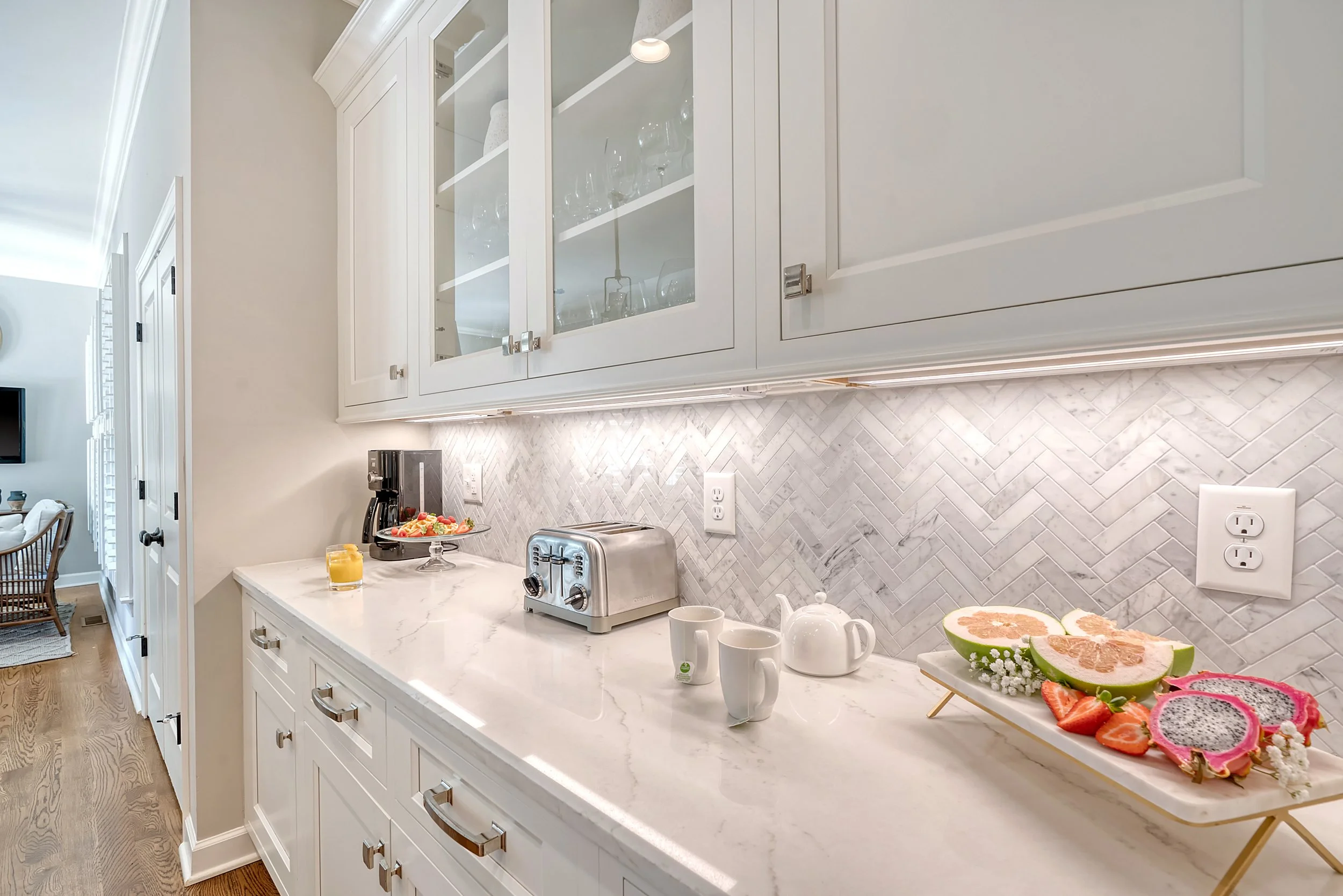 Kitchen countertop with fruit, teapot, mugs, toaster, coffee maker, and decorations, with white upper cabinets and a marble backsplash.