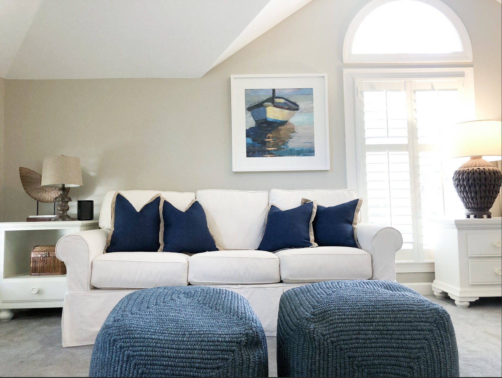 Living room with white sofa, five navy blue pillows, two blue textured ottomans, window with shutters, painting of a boat on water, table lamps, and decorative items.