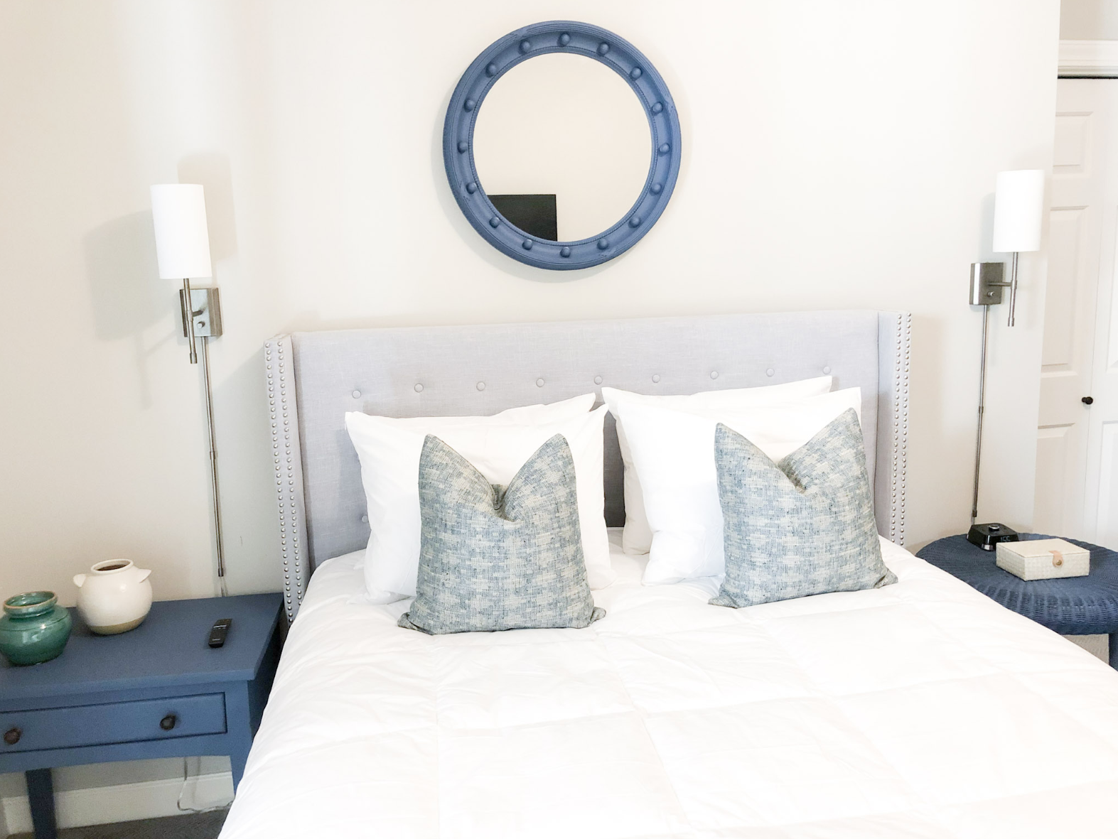 A tidy bedroom with a bed featuring a light gray upholstered headboard. There are two pillows with gray decorative pillow covers, two white pillows behind them, and a white comforter. On either side of the bed are wall-mounted lamps with white shades