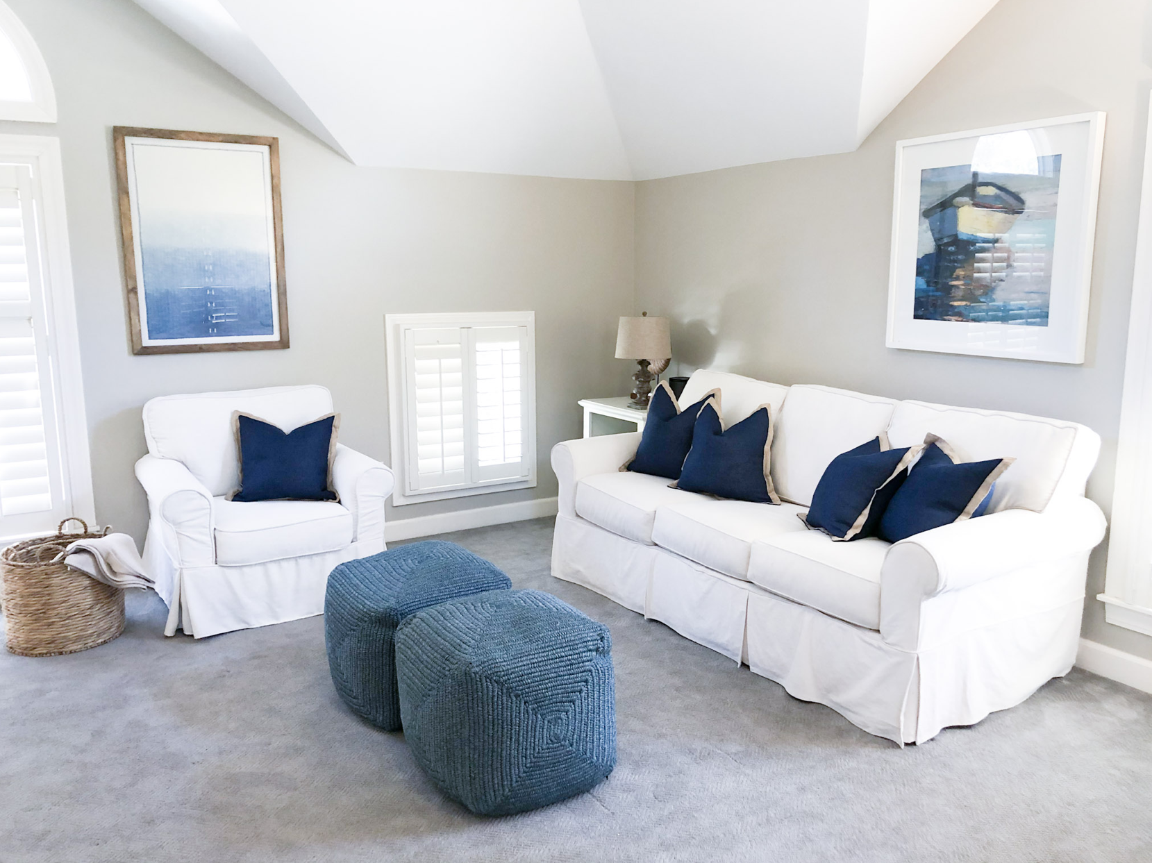 Living room with white sofa and armchair, blue pillows, two blue ottomans, a side table with a lamp, framed abstract art on light-colored walls, and a basket with blankets.
