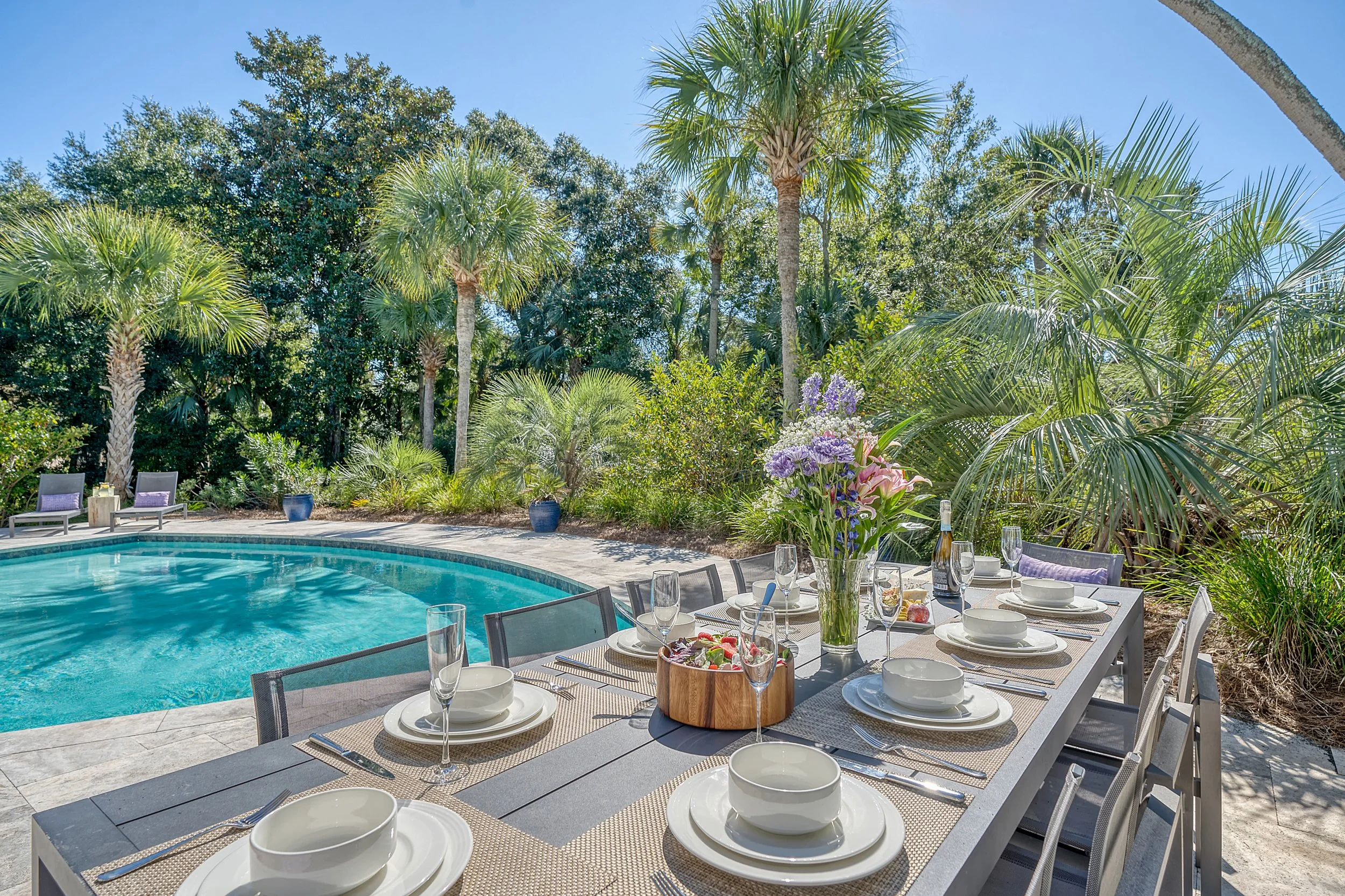Outdoor dining table set with white dishes, glasses, and a floral centerpiece, next to a large swimming pool surrounded by tropical palm trees and greenery on a sunny day.