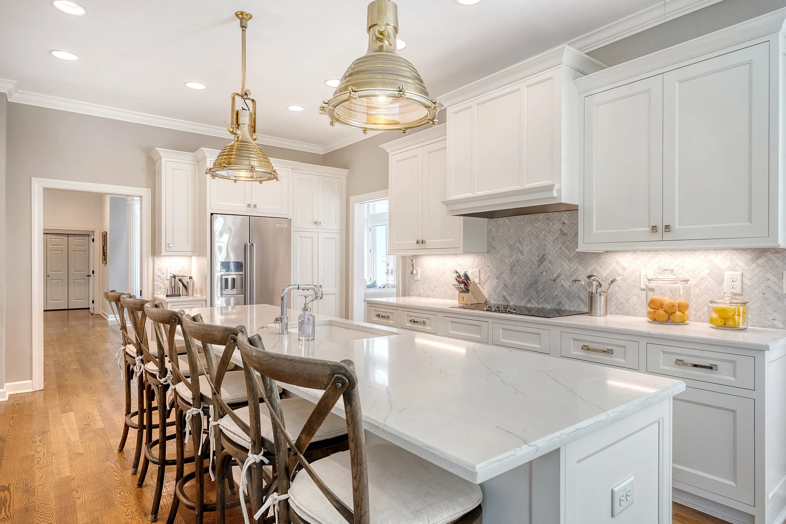 Modern white kitchen with marble island, wooden bar stools, and brass pendant lights.