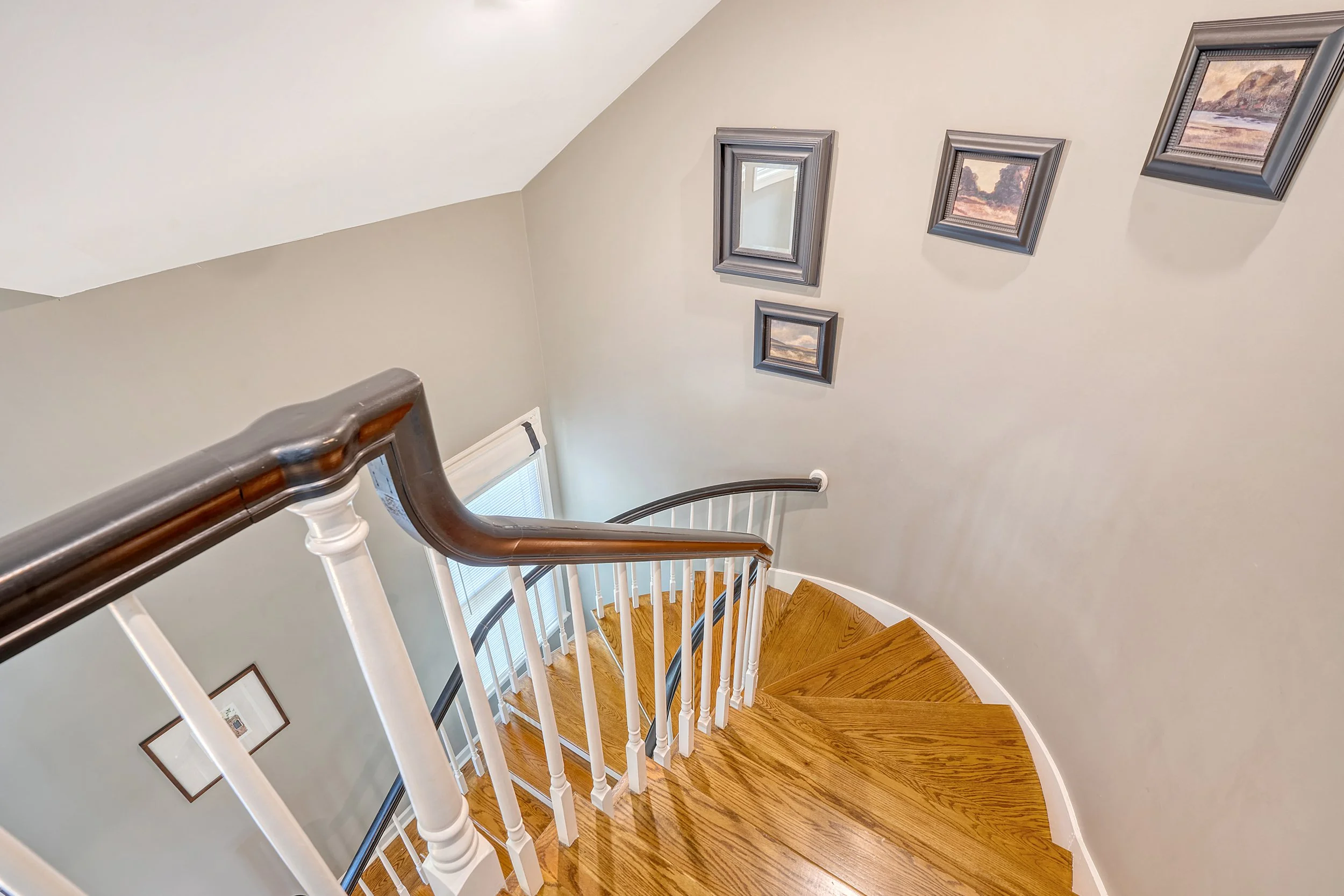 View of a curved wooden staircase with white balusters and a dark wood handrail in a home, with framed landscape pictures on a light beige wall.