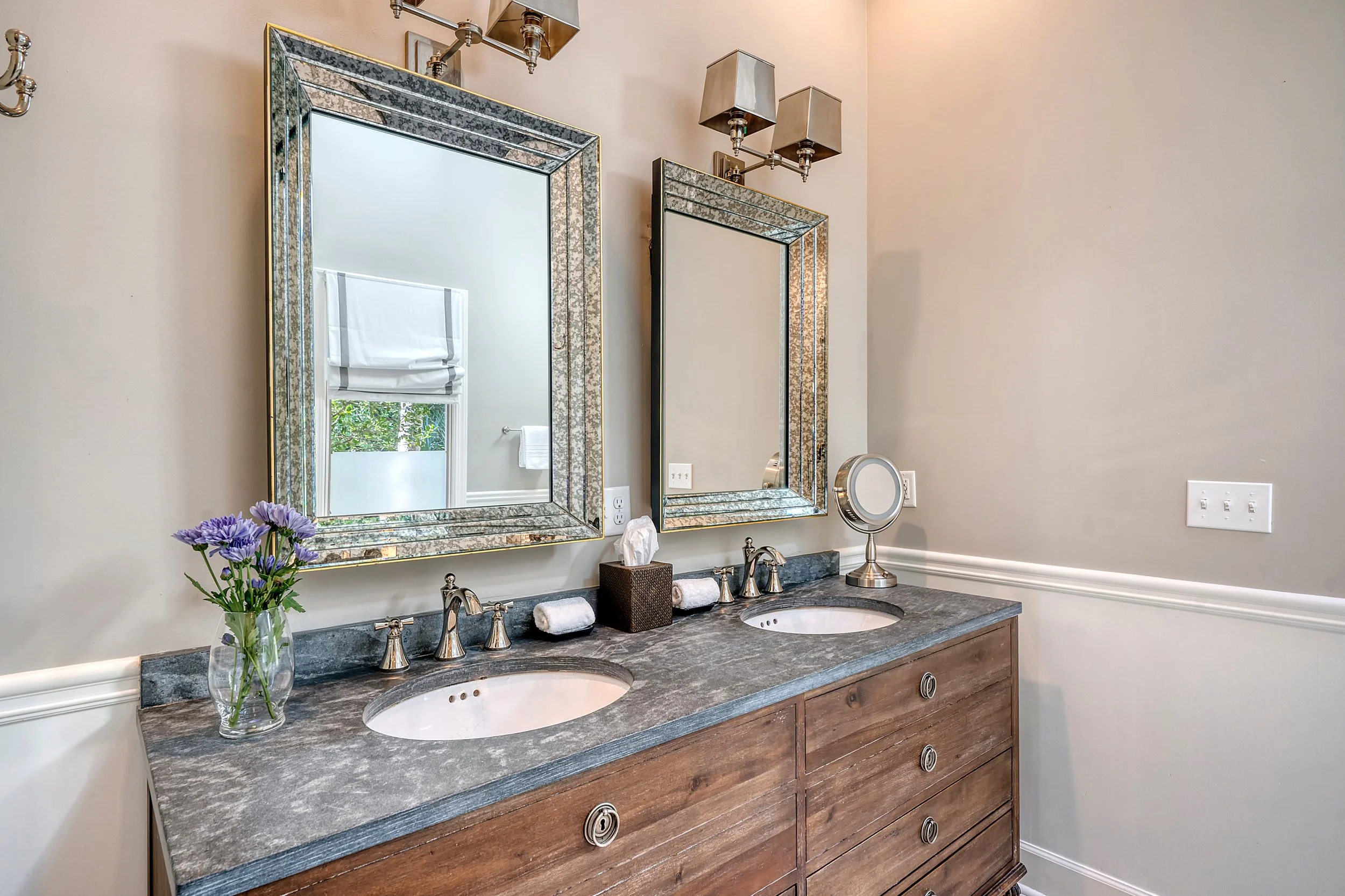 A bathroom double vanity with two mirrors, a granite countertop, and a wooden cabinet below. Decor includes a vase of purple flowers, a tissue box, a small mirror, and rolled towels. The wall has a light switch and a power outlet.