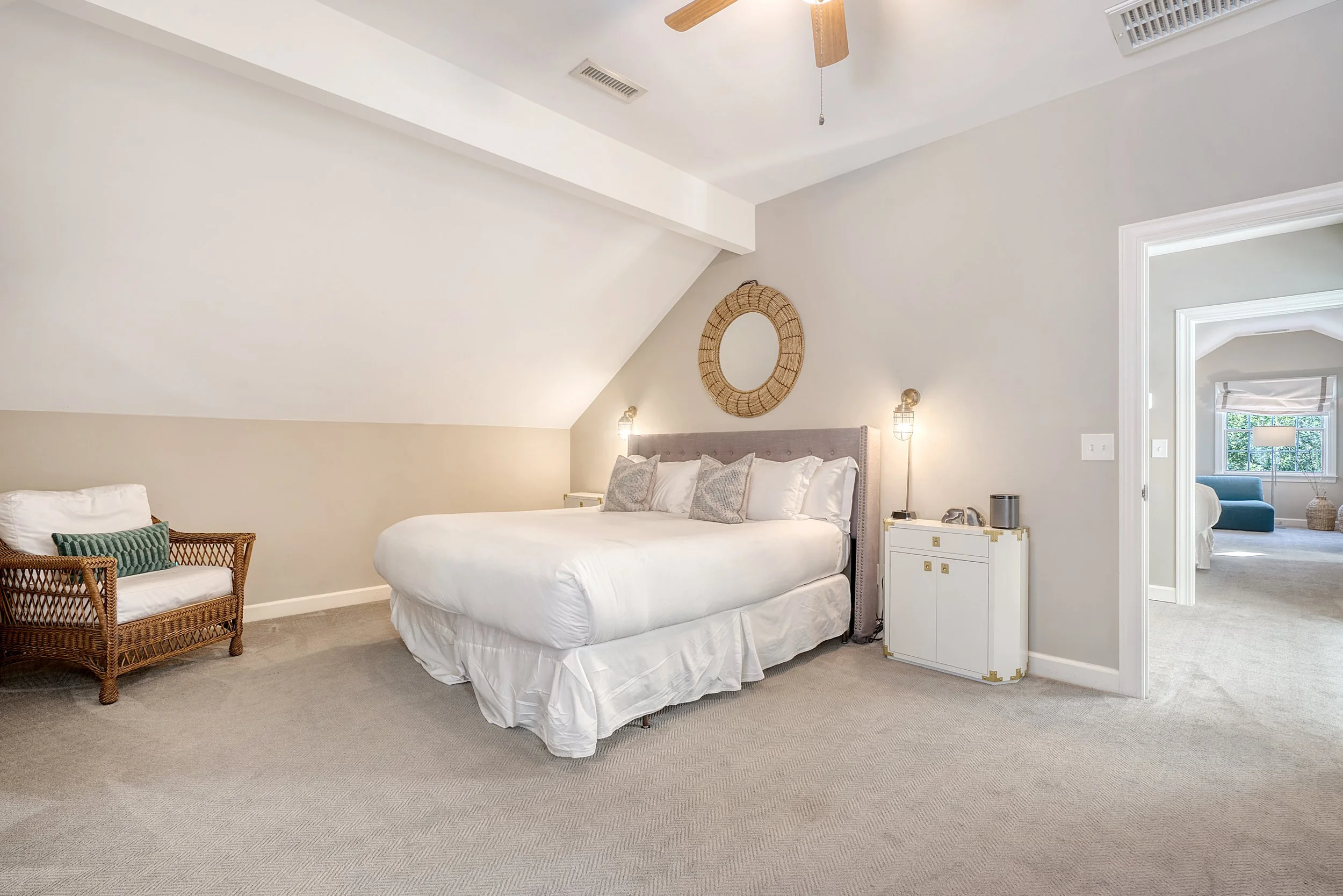 A bedroom with a sloped ceiling, a bed with white bedding, a wicker chair with a green pillow, and a small white nightstand with lamps and decor. There is a circular mirror on the wall above the bed and a doorway leading to another room with a view o