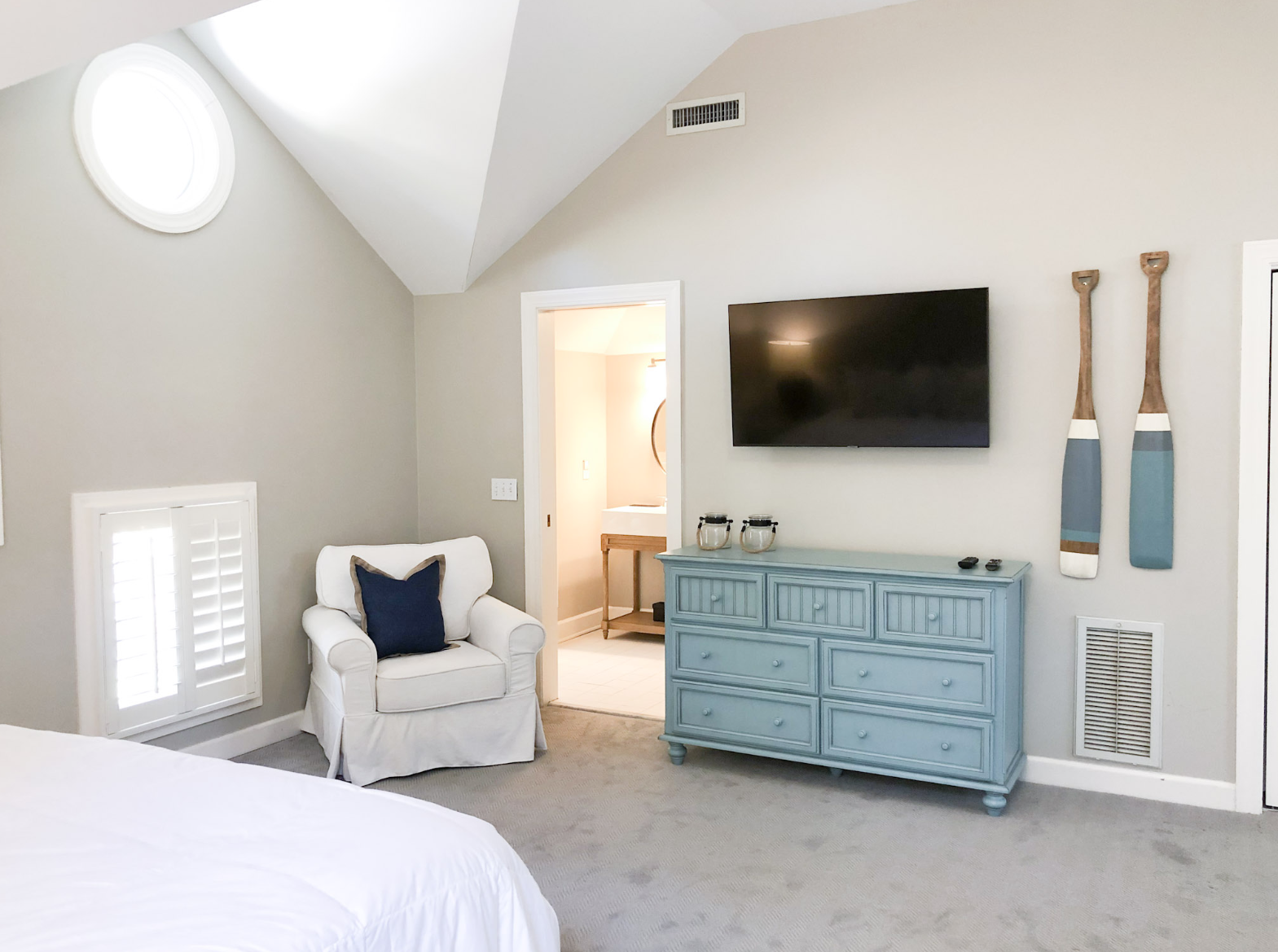 A cozy bedroom with beige walls, a white armchair with a navy pillow, a blue dresser, a mounted flat-screen TV, and decorative oars on the wall.