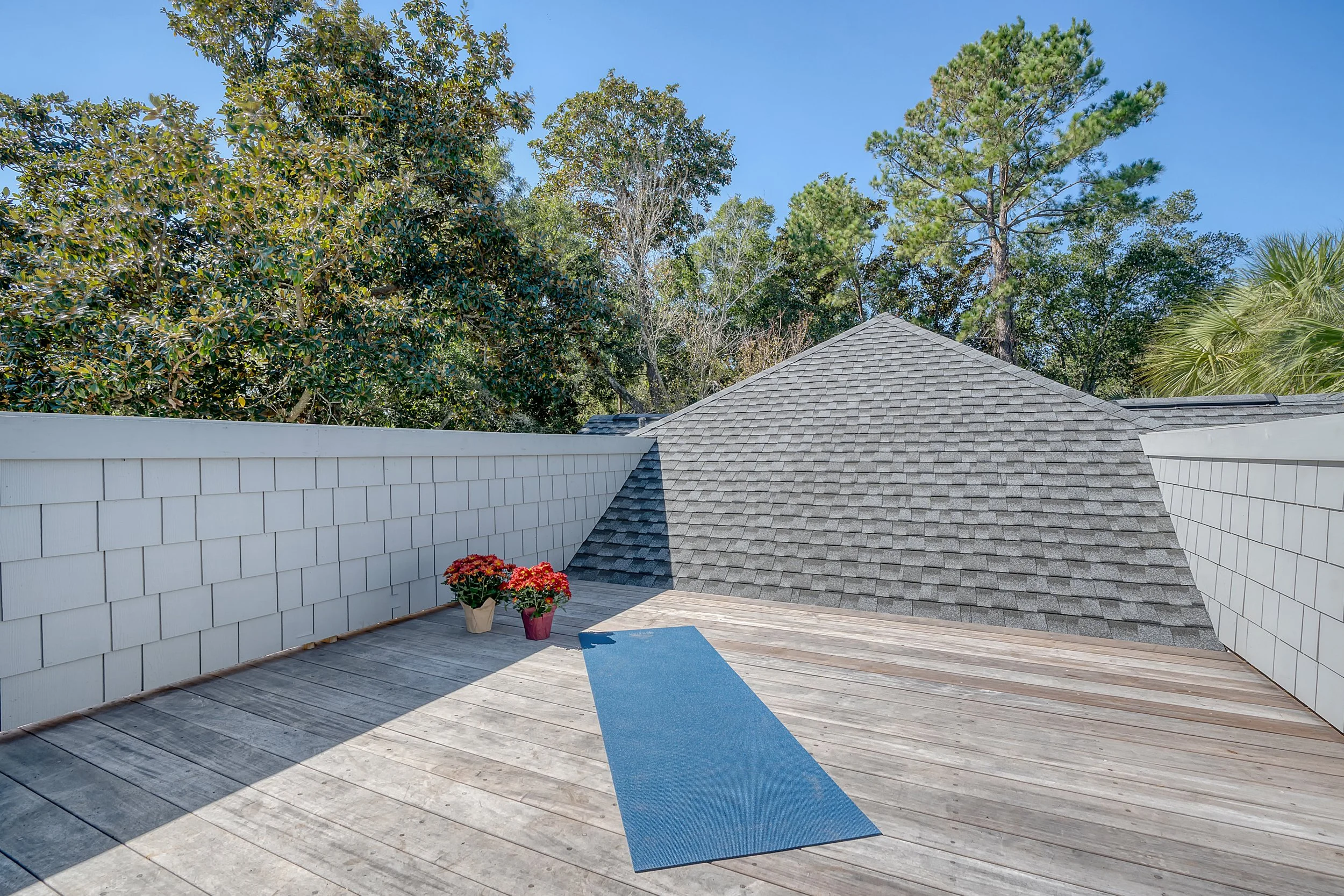 Rooftop deck with a blue yoga mat and two potted plants, surrounded by trees and white railing, with a clear blue sky.